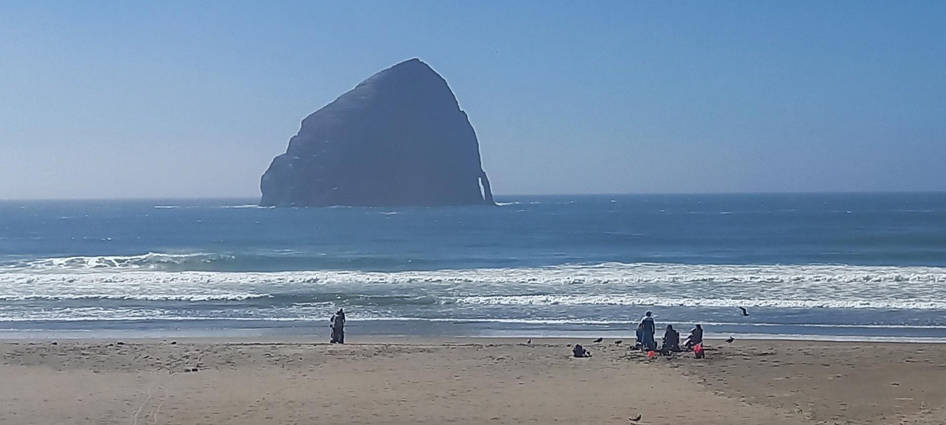 Large sea stack in ocean, people on sandy beach with waves, bright day.