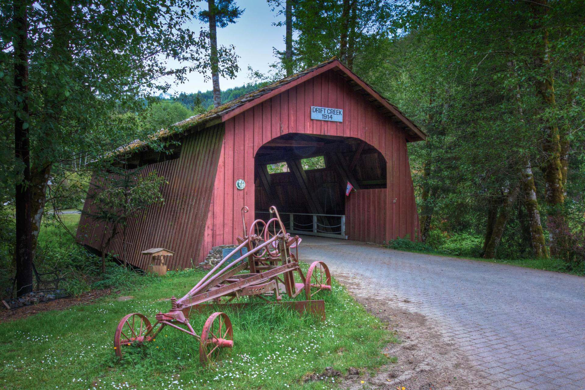 Rustic red covered bridge with old farm equipment in a lush forest.