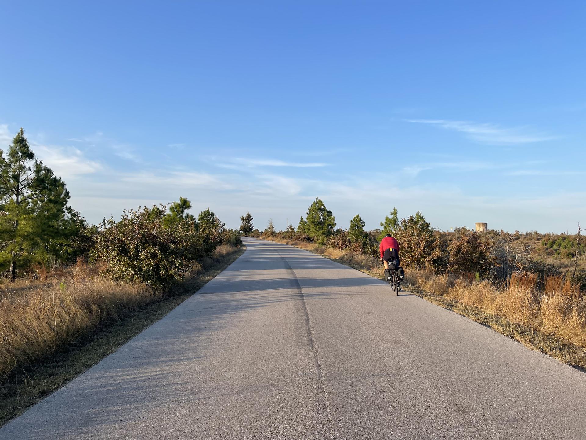A cyclist rides on a straight road through a dry, open landscape.