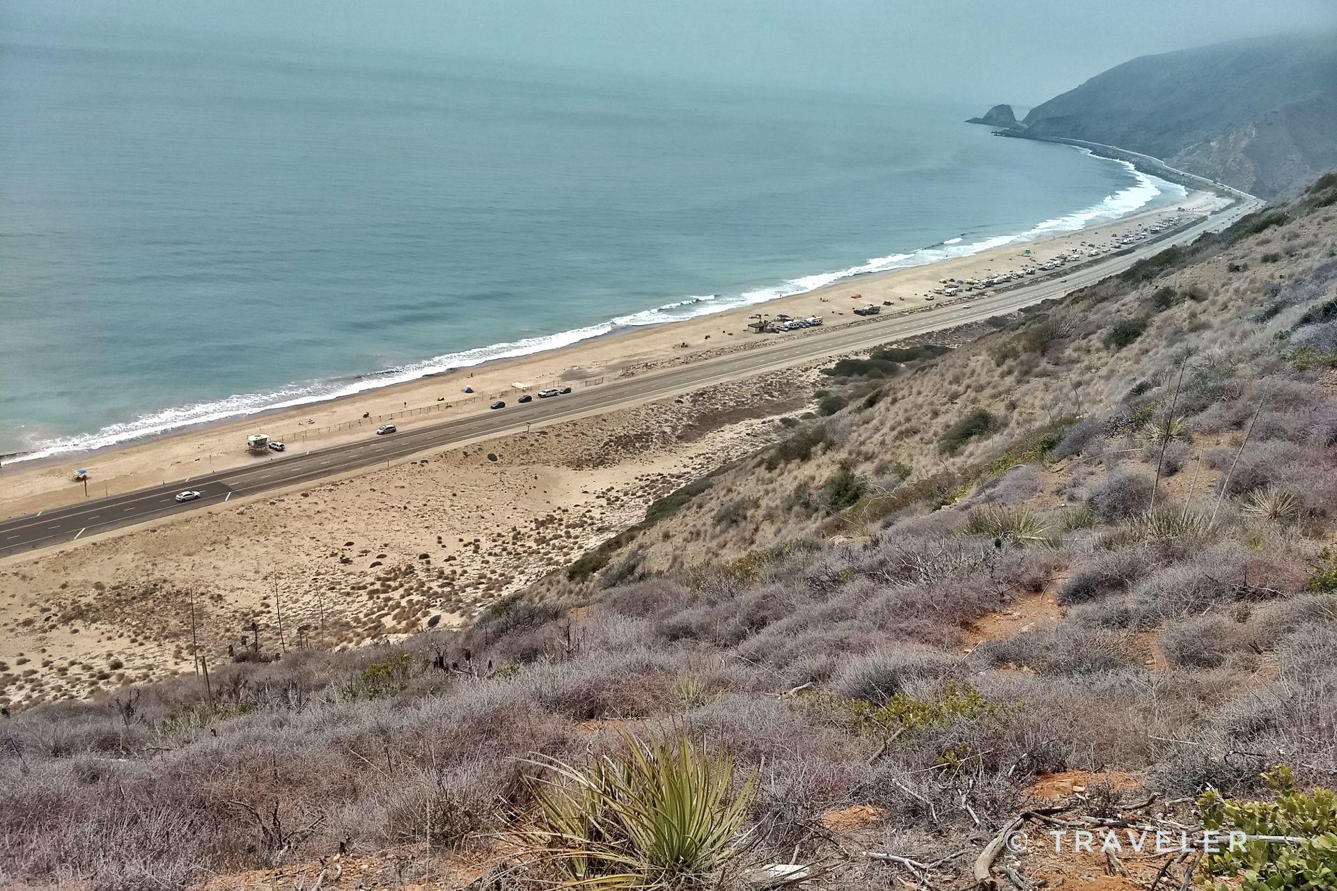 Overcast view of a coastal highway beside a sandy beach and ocean.