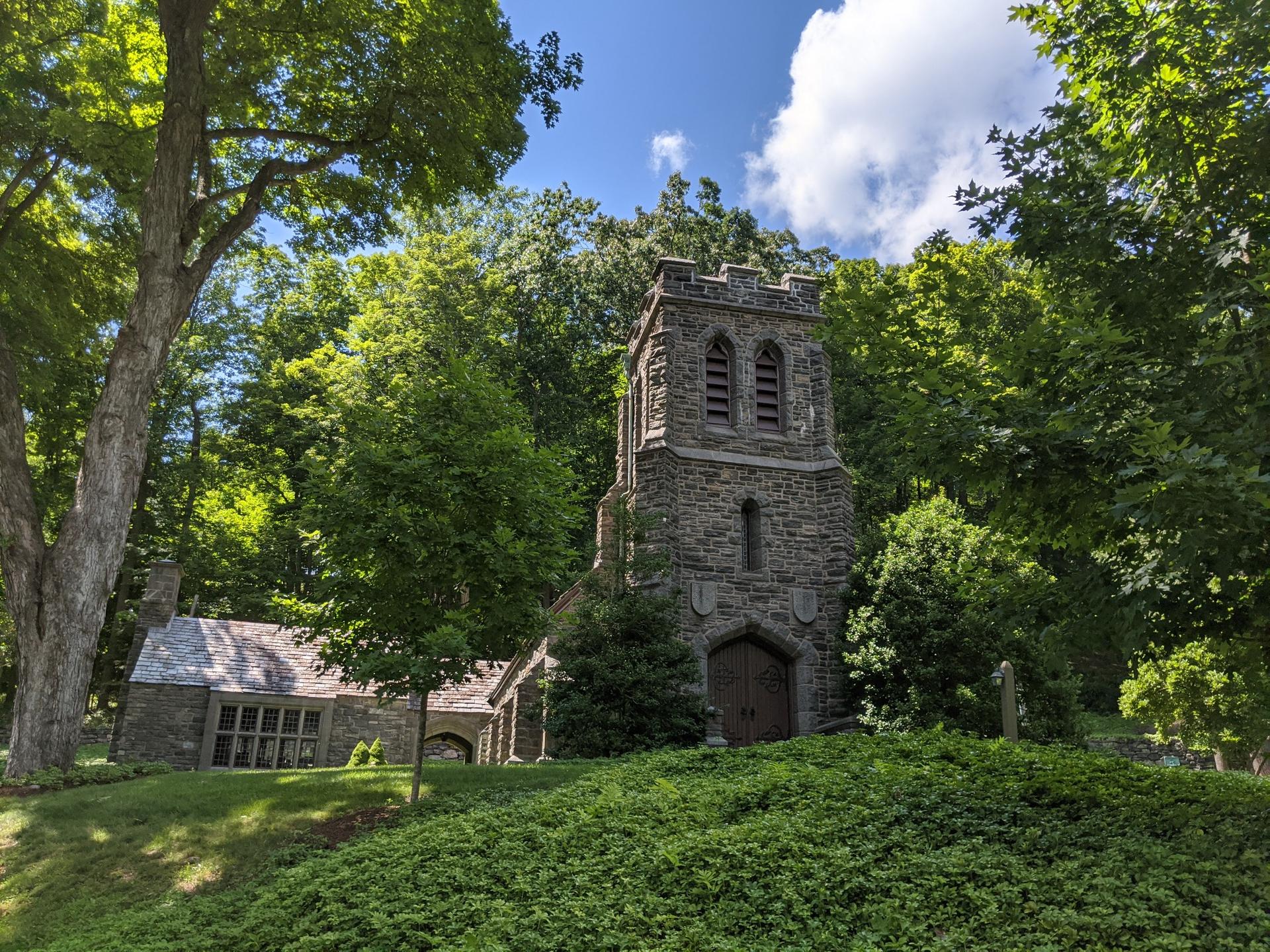 An old stone church tower nestled among dense green trees on a sunny day.