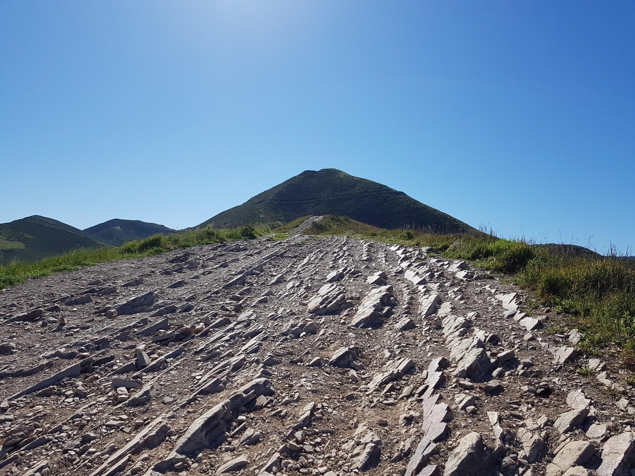 Rocky path ascends towards a distant mountain under a bright, clear sky.
