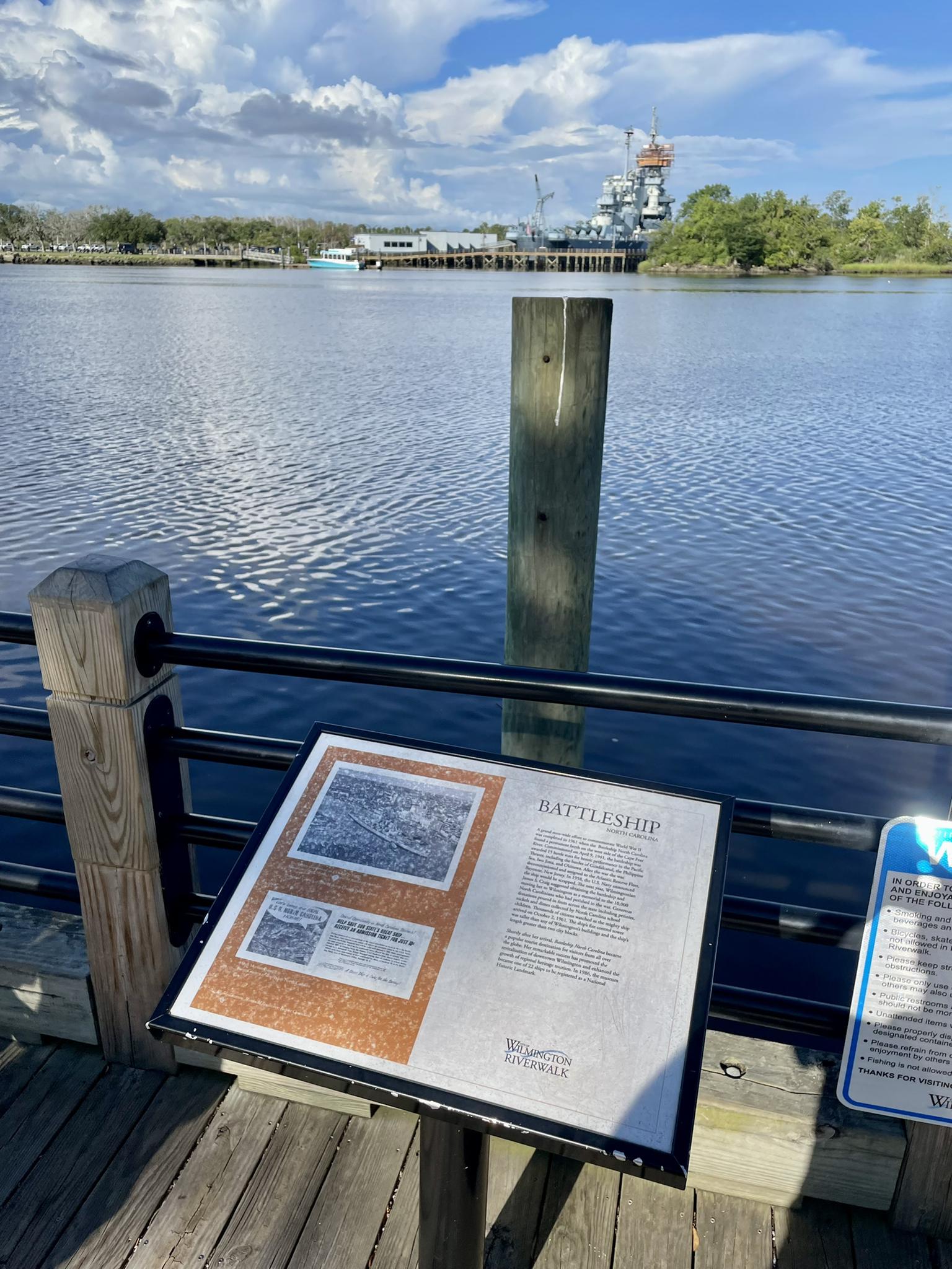 Informational sign on a pier overlooking a river with a distant warship.