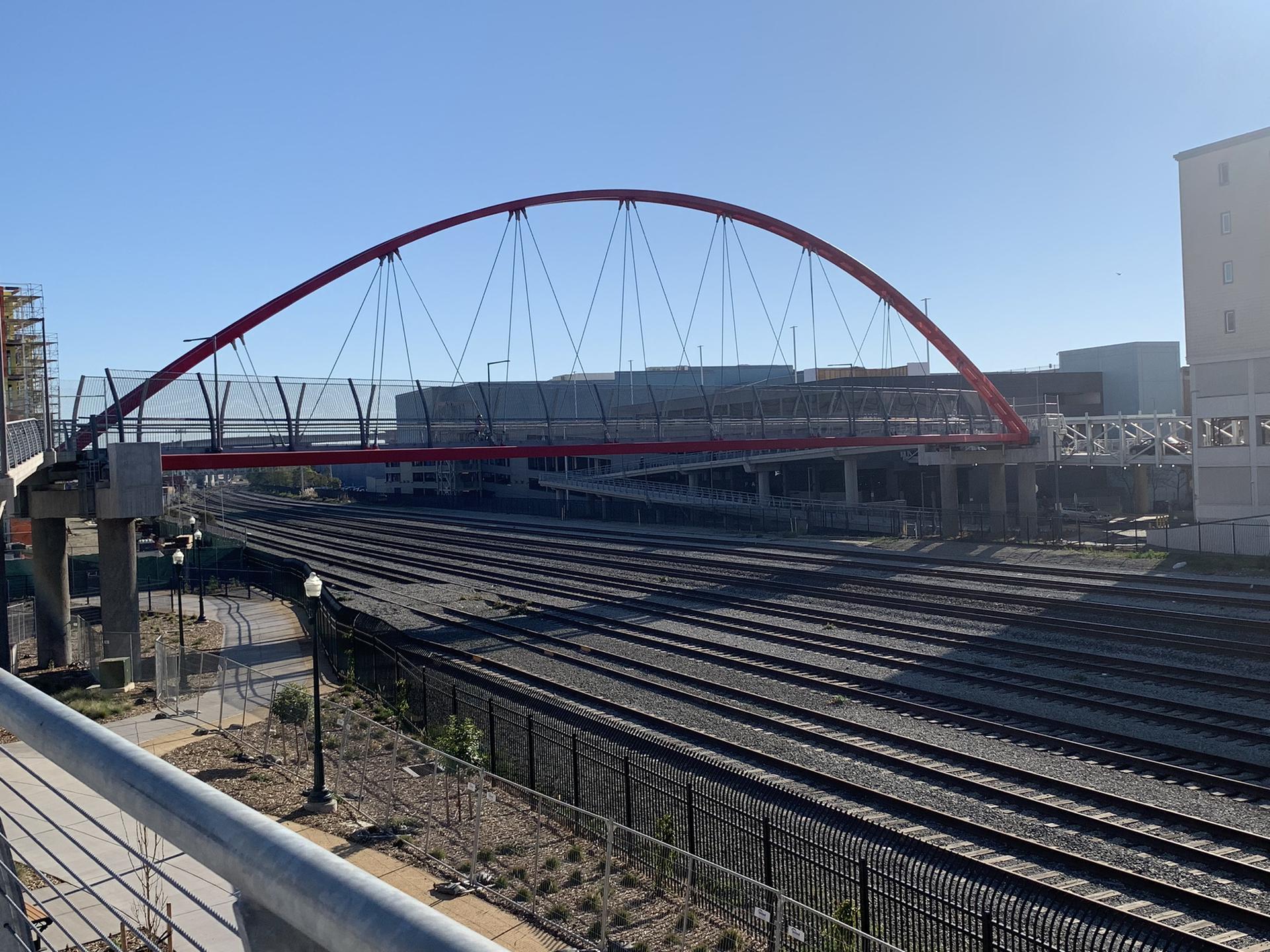 Red arched pedestrian bridge spans over numerous train tracks on a sunny day.
