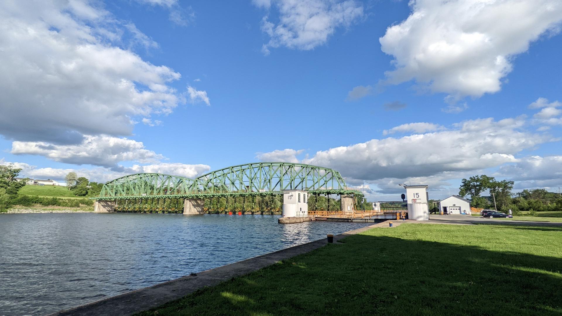 A large green truss bridge spans a river under a bright, cloudy sky.