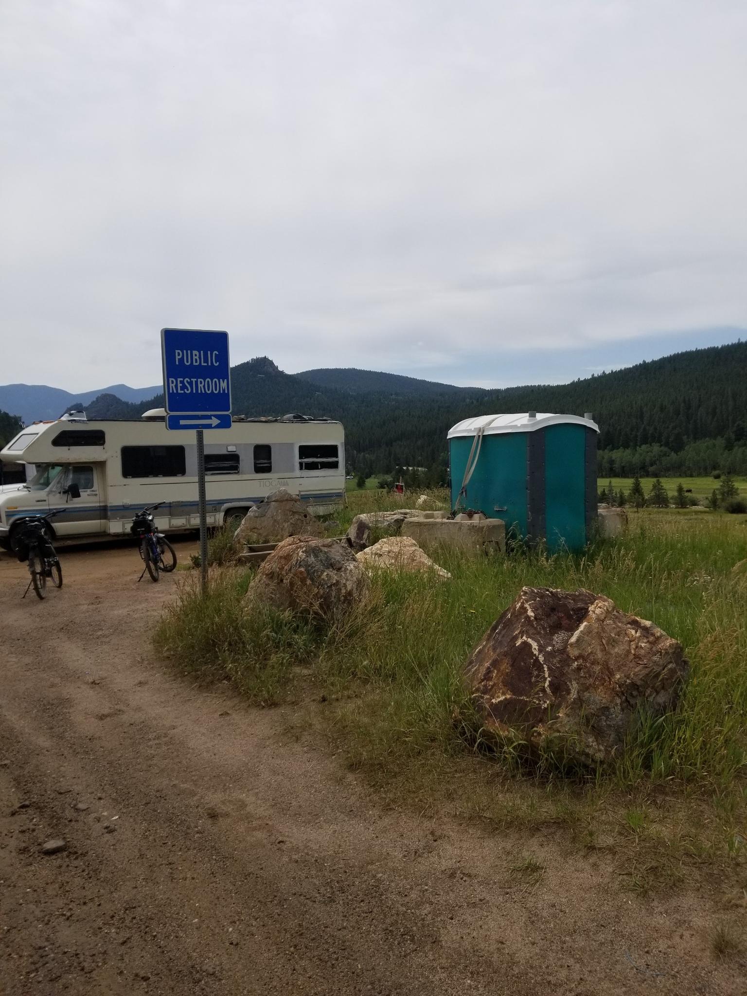 Dirt road with public restroom sign, RV, and portable toilet under overcast sky.