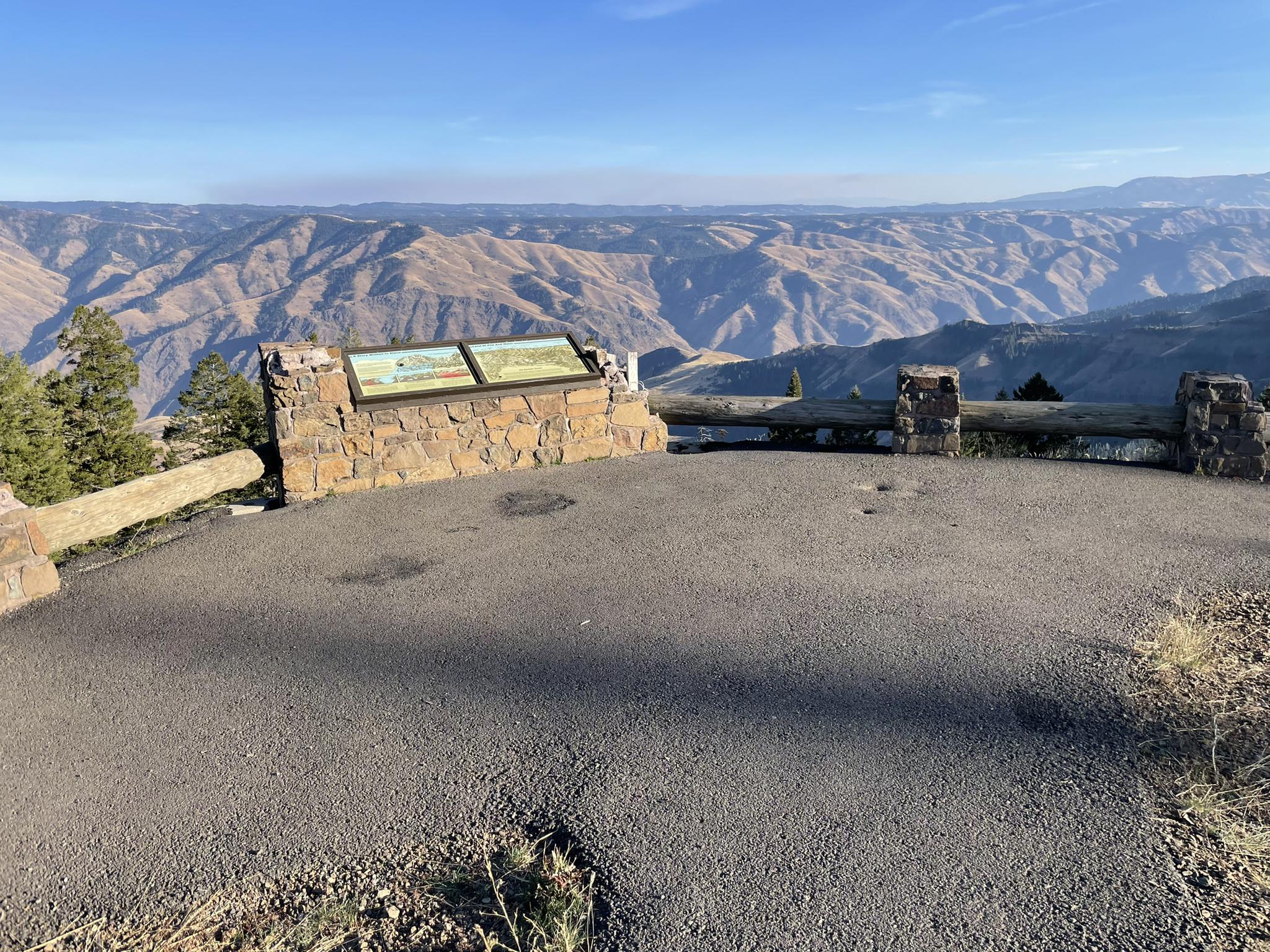 Paved overlook with stone sign and fence, vast rugged mountains beyond.