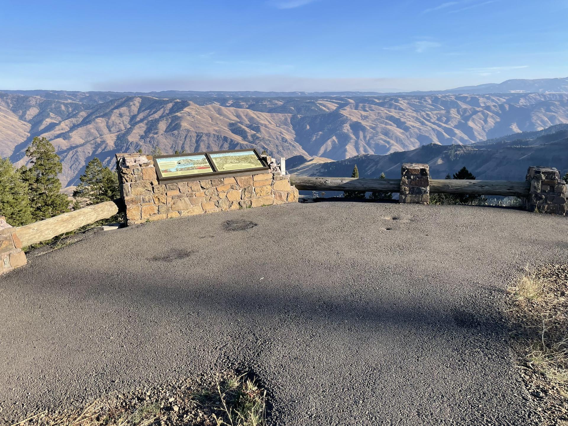 Paved overlook with stone sign and fence, vast rugged mountains beyond.