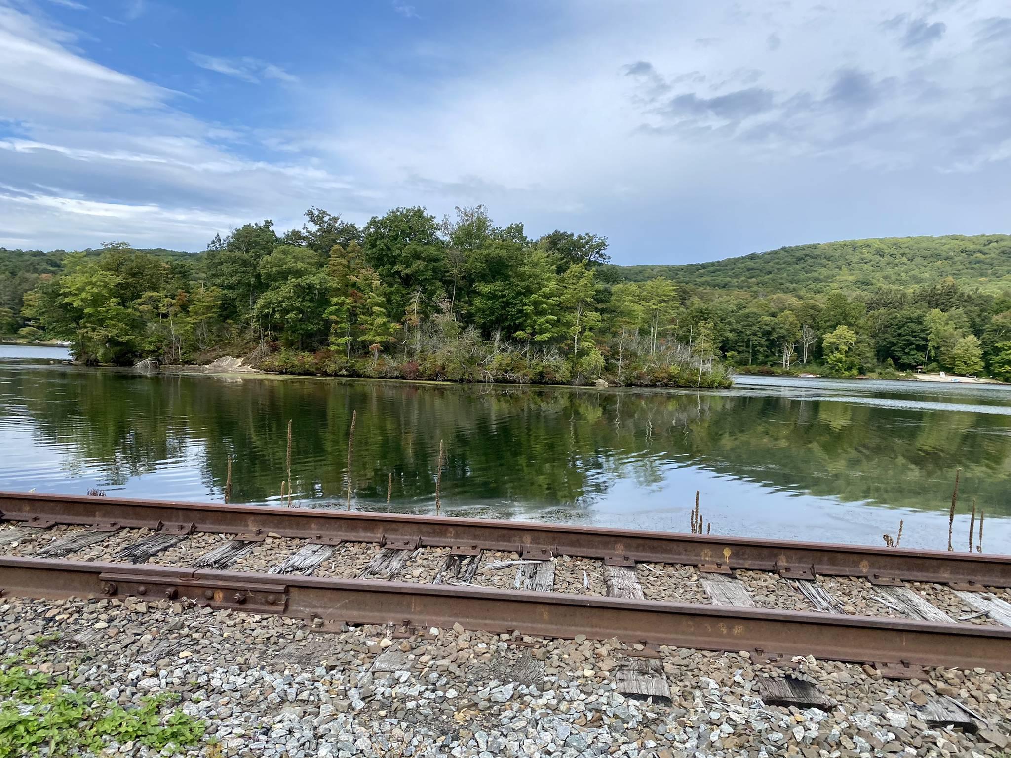 Calm lake with forested islands next to old railroad tracks under cloudy sky.