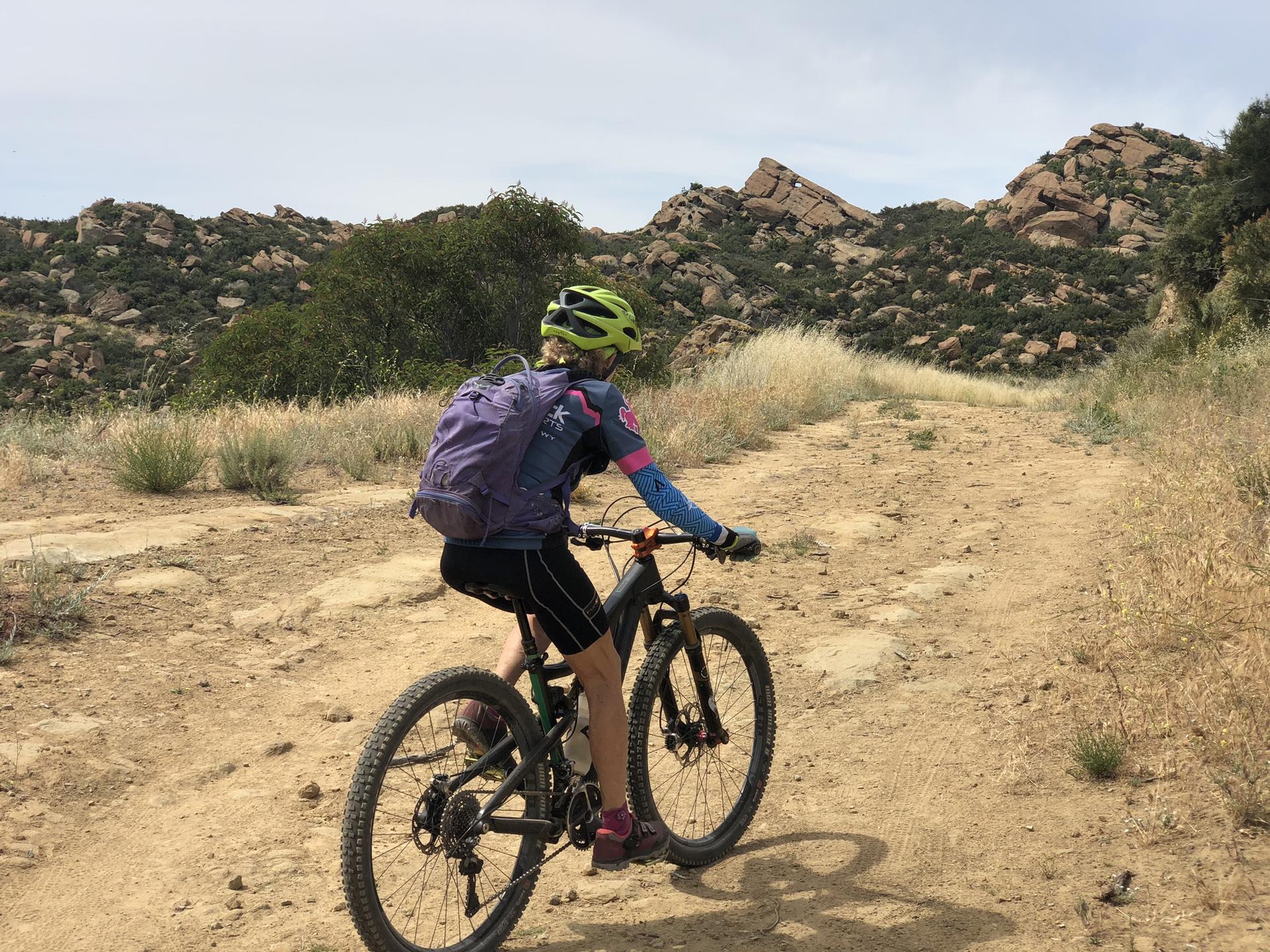 Cyclist on a dirt path, wearing a helmet and backpack, with rocky hills.