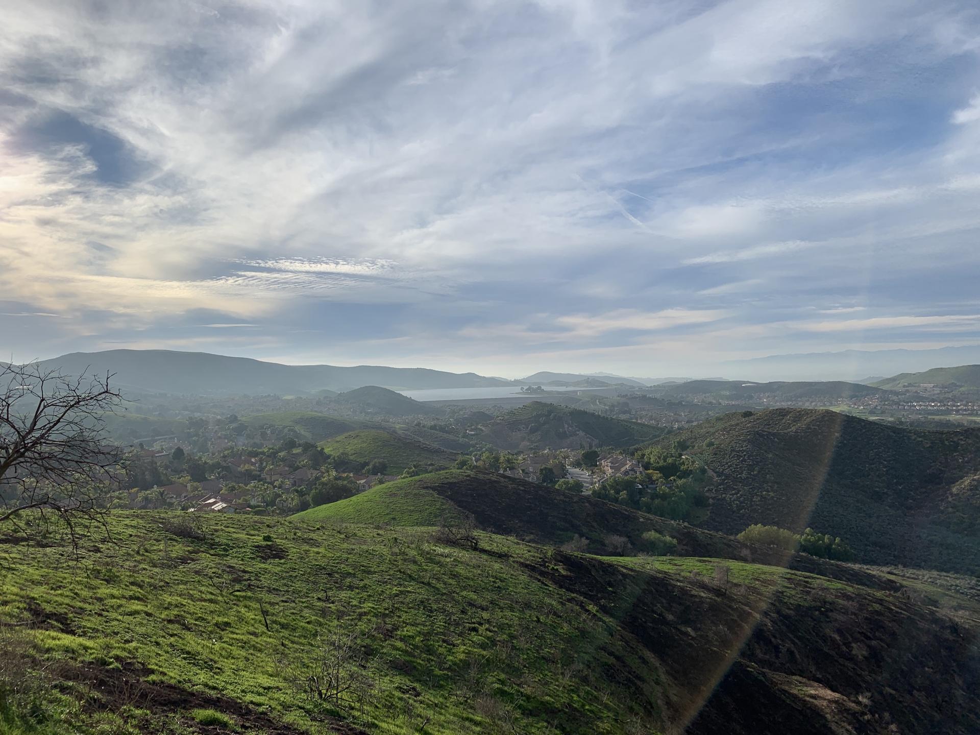 Expansive view of green, hazy hills and distant water under a cloudy sky.