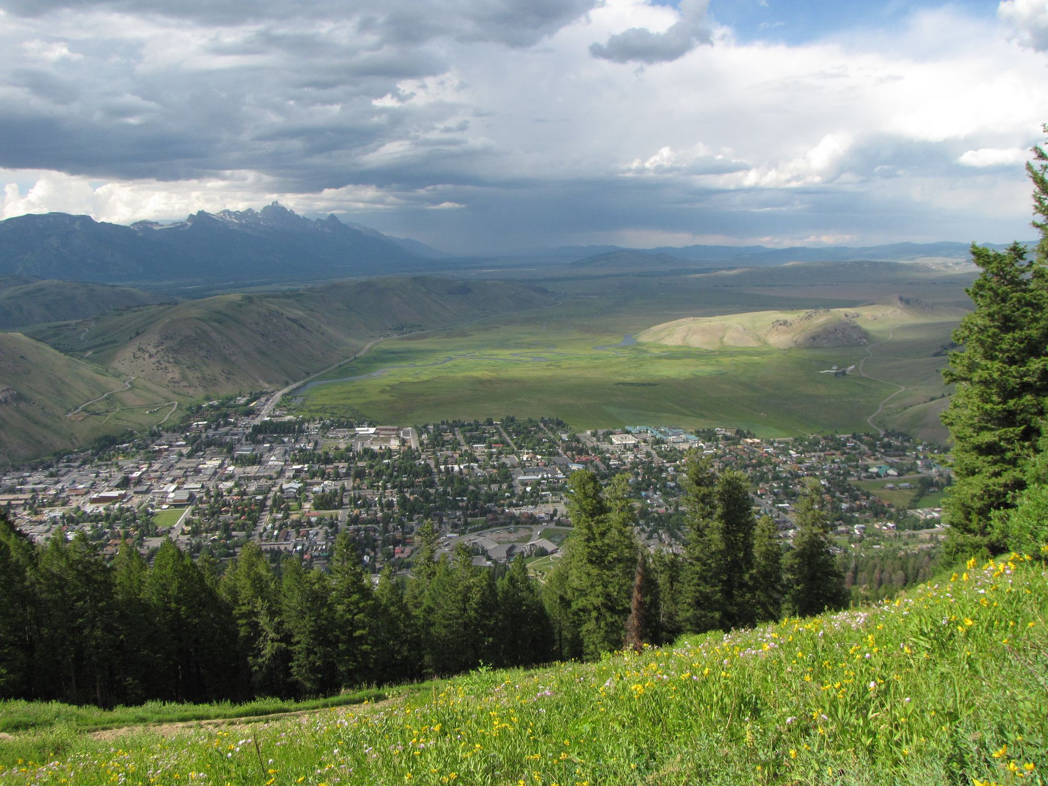 Overhead view of a town in a valley, green hills, and distant mountains under clouds. - Snow King Mountain Park – Snow King Mountain loop from Jackson