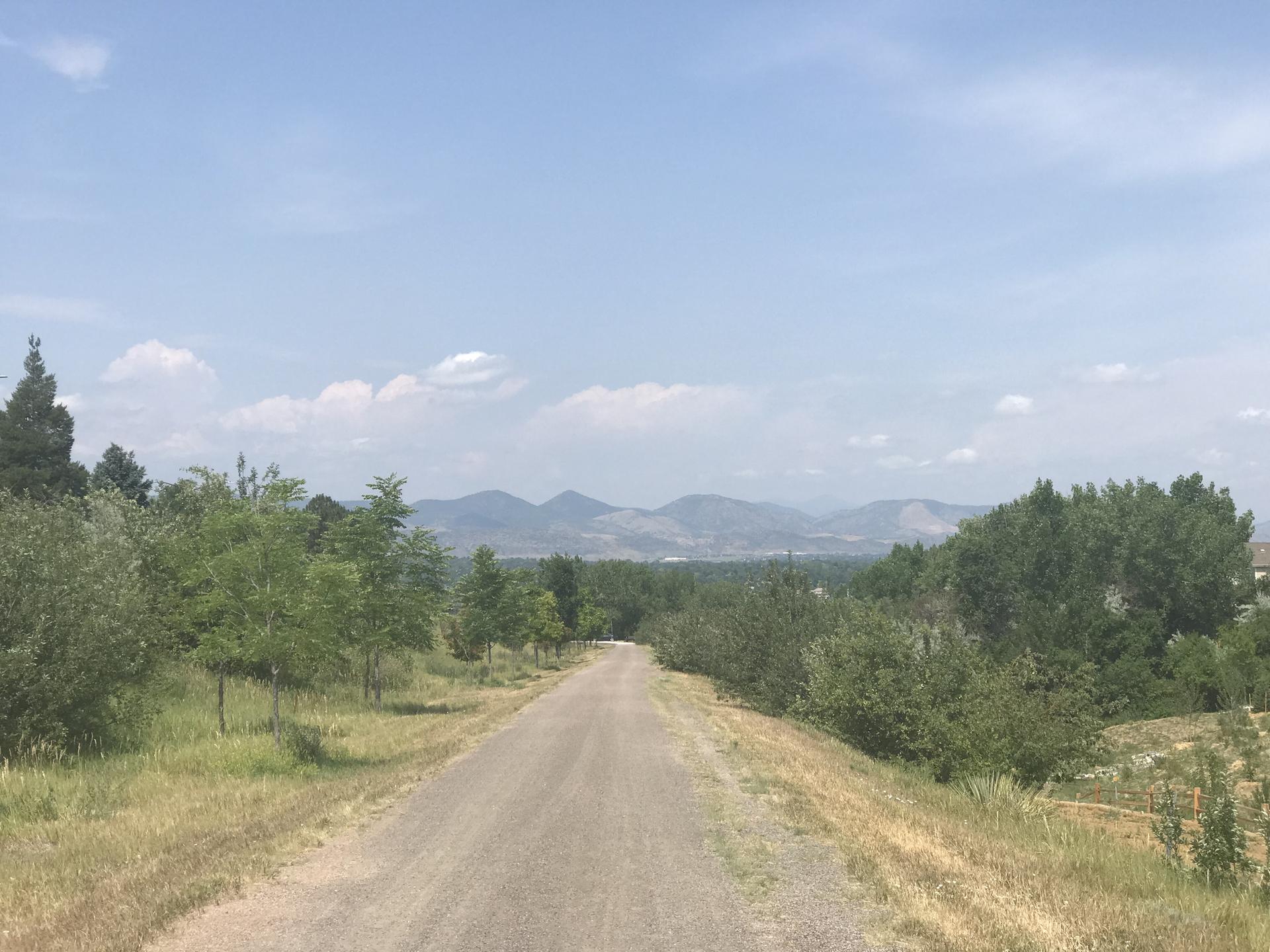 Dirt path through green trees, leading to distant mountains under a blue sky.