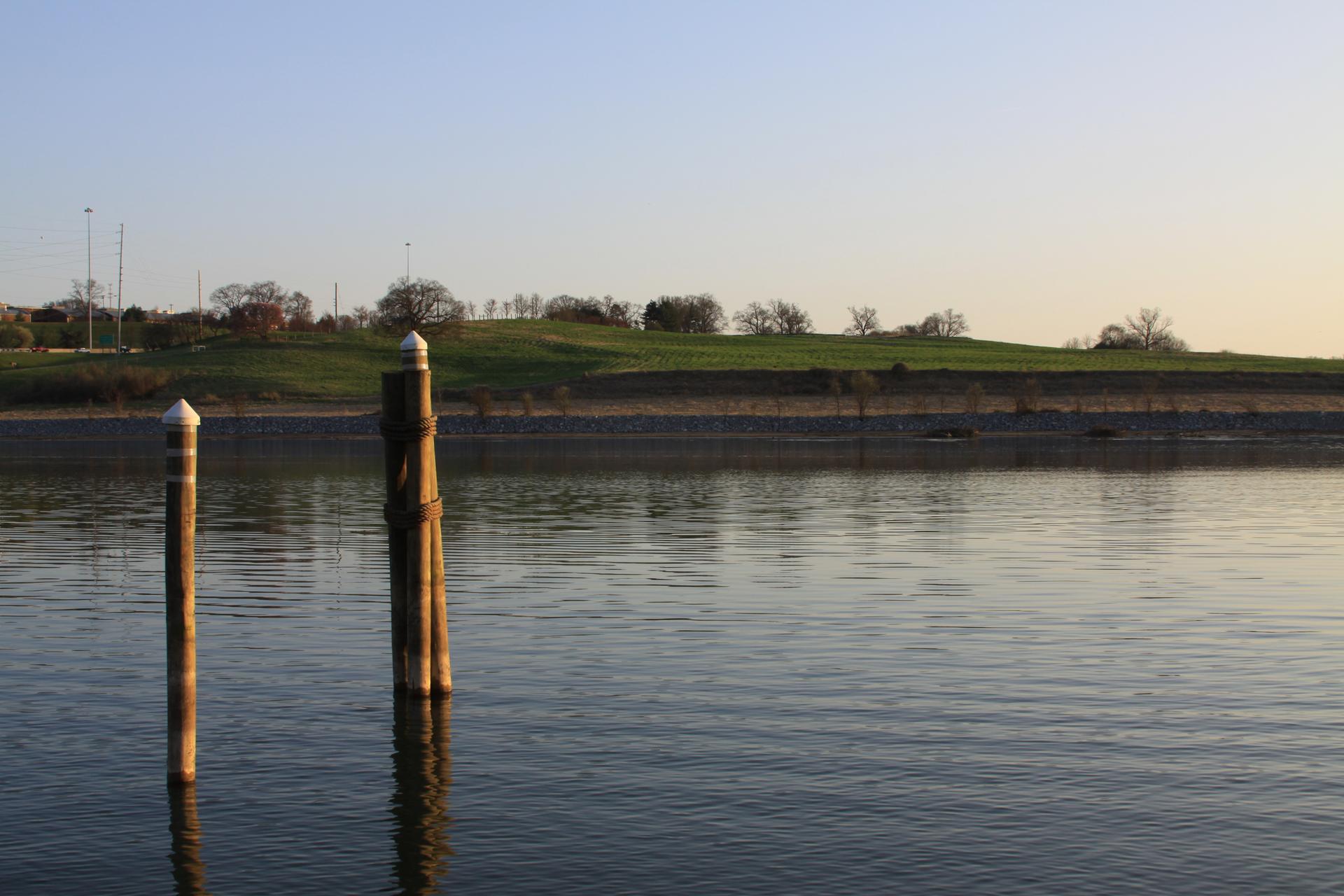 Two wooden posts in calm water with a grassy hill under soft light.