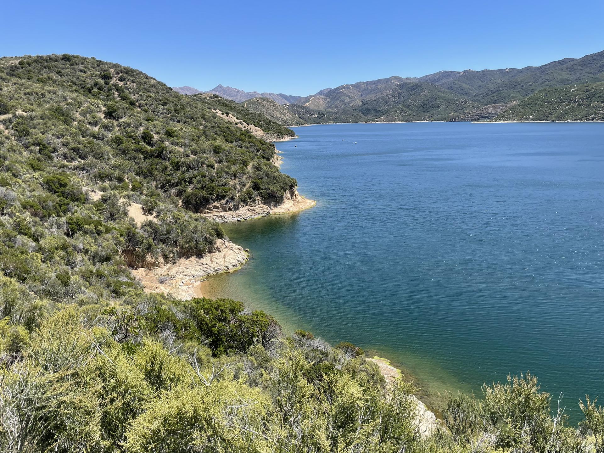 Expansive blue lake bordered by steep, brush-covered hills under a bright sky.