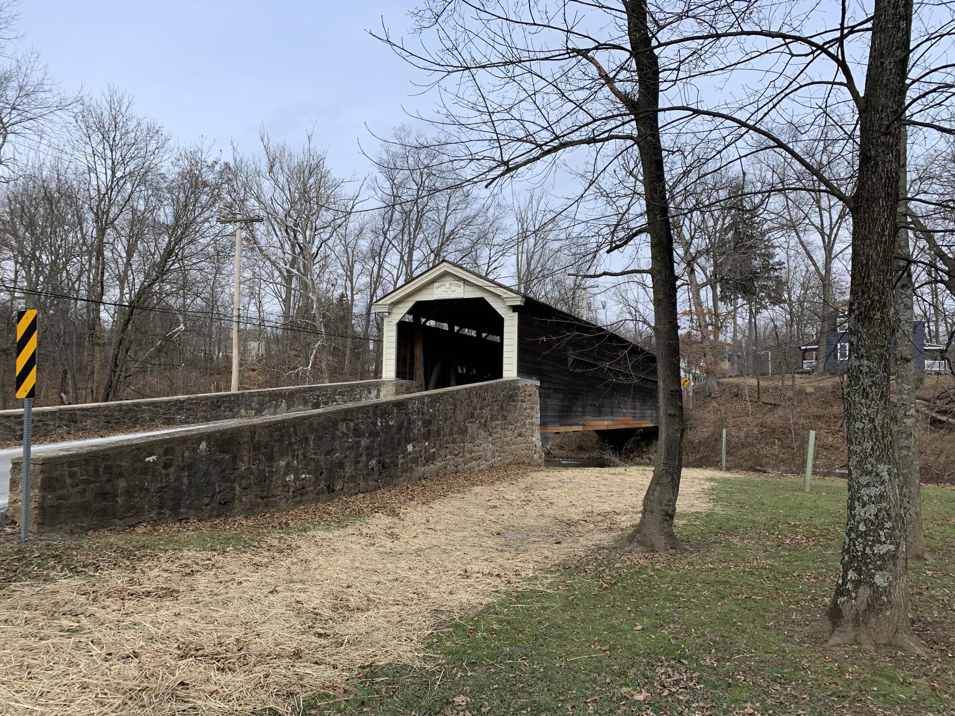 A covered wooden bridge with a stone base, surrounded by bare trees under an overcast sky.
