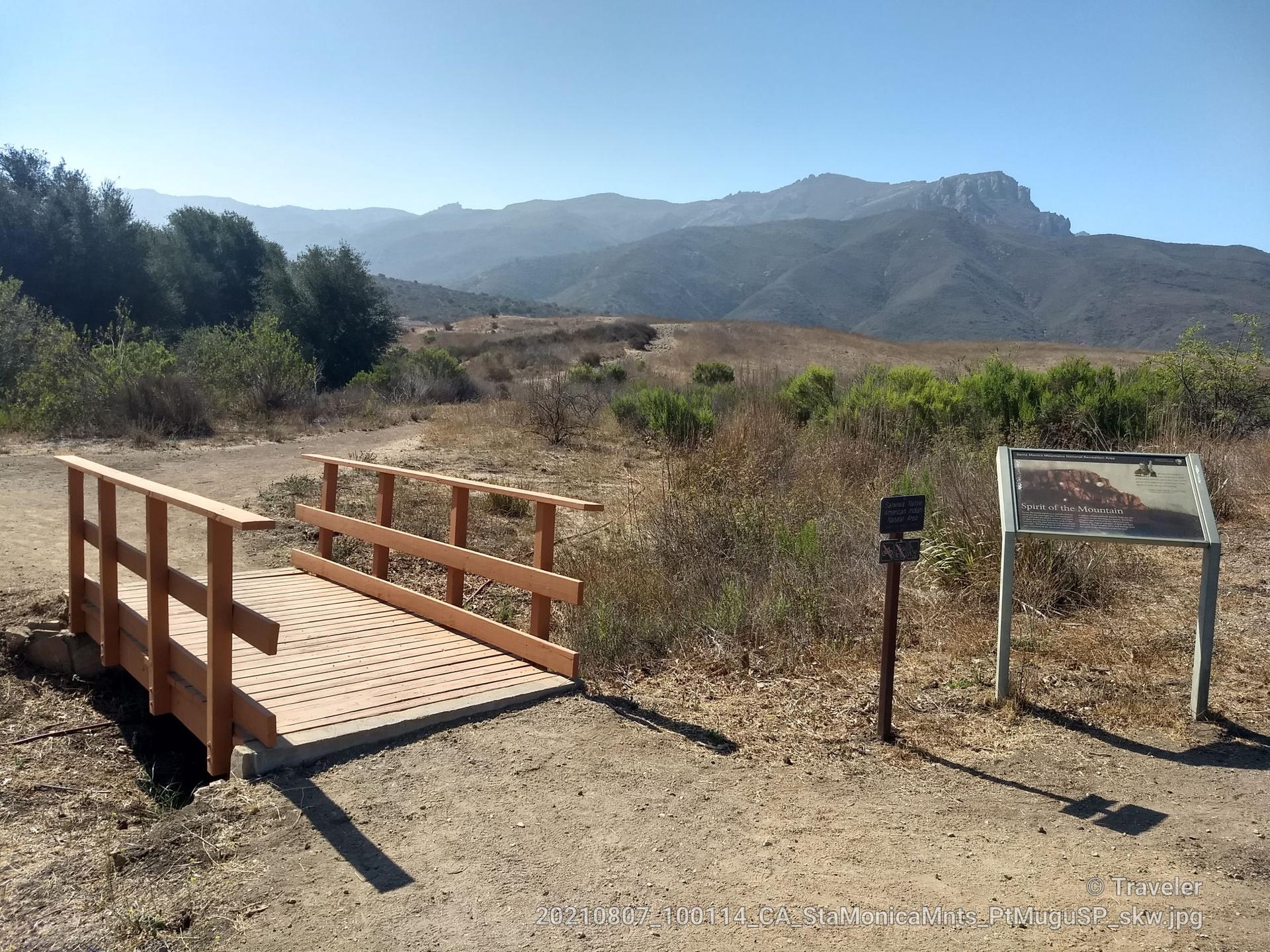 Wooden footbridge and information sign in a dry field with distant mountains.