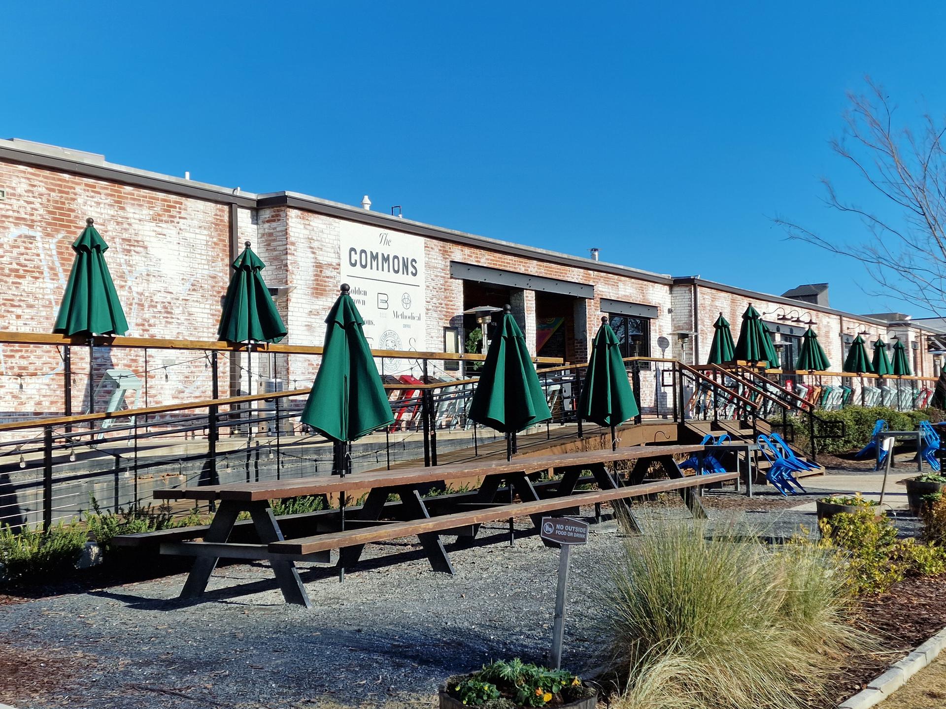 Sunny outdoor patio with green umbrellas and picnic tables next to a brick building.