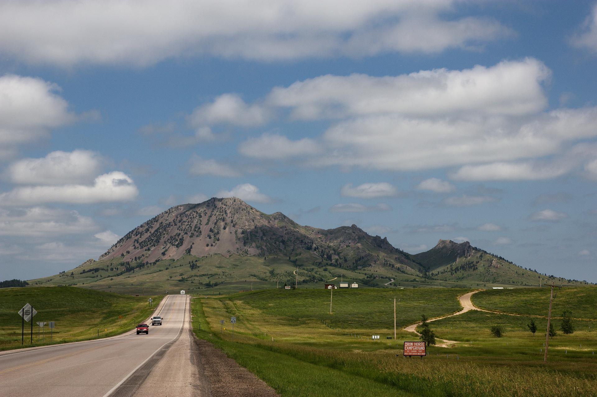 A highway leads to large, green, rocky mountains under a bright, cloudy sky.