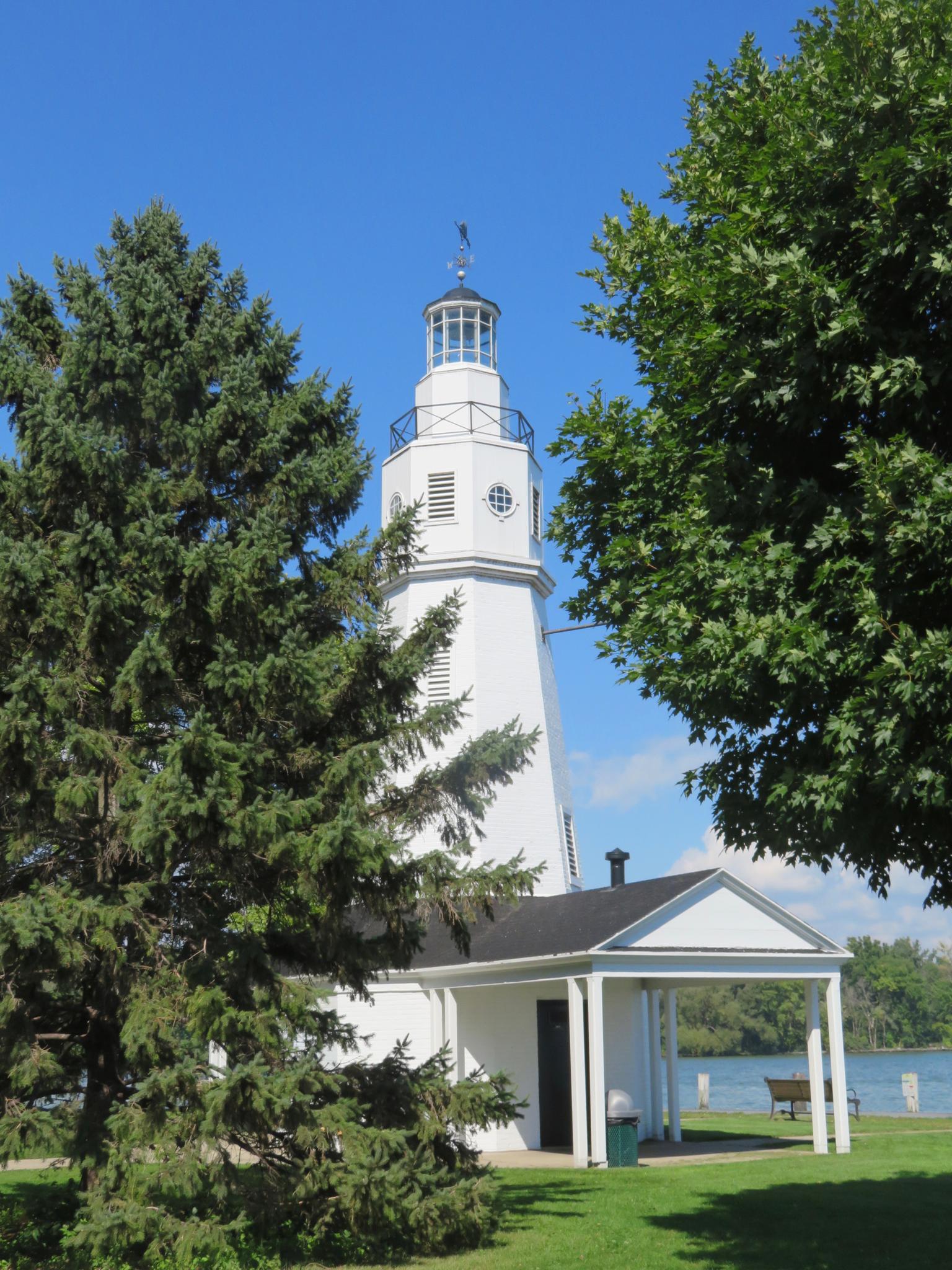 A white lighthouse building stands between green trees under a blue sky.