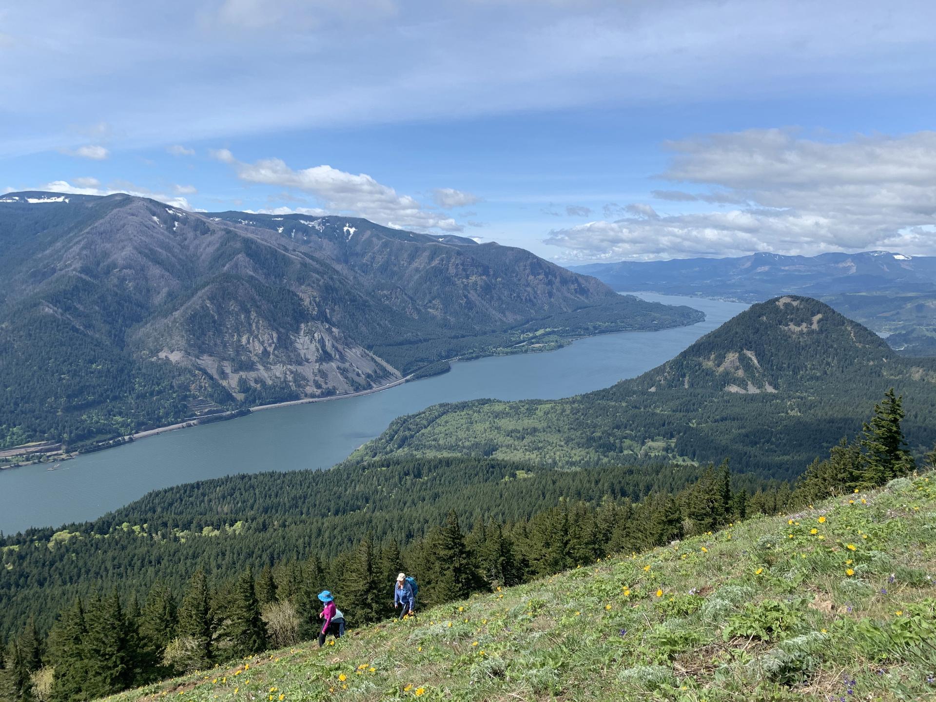 Two hikers ascend a grassy mountain slope overlooking a wide river.