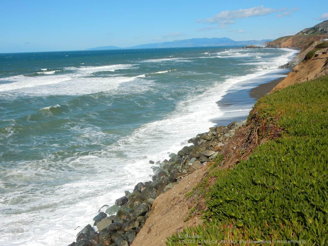 Powerful ocean waves crash onto a rocky beach below a green cliff.