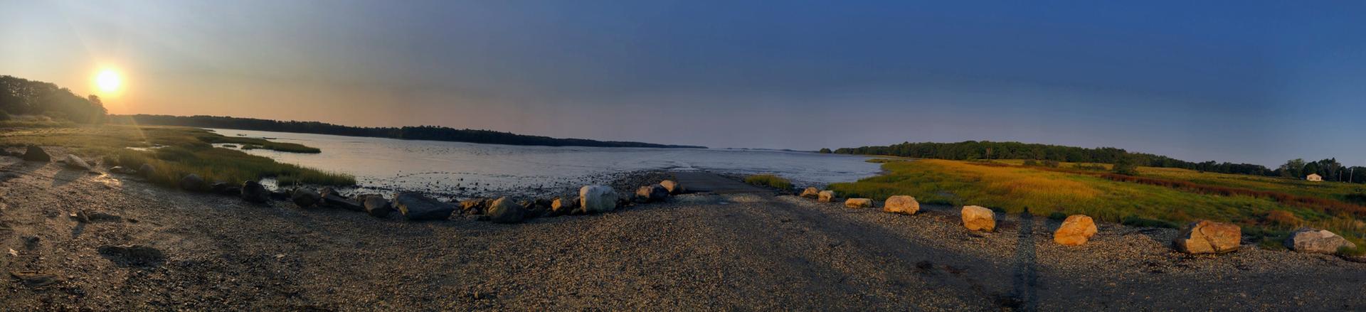 Panoramic sunset over a calm bay with sandy shore and grassy banks.