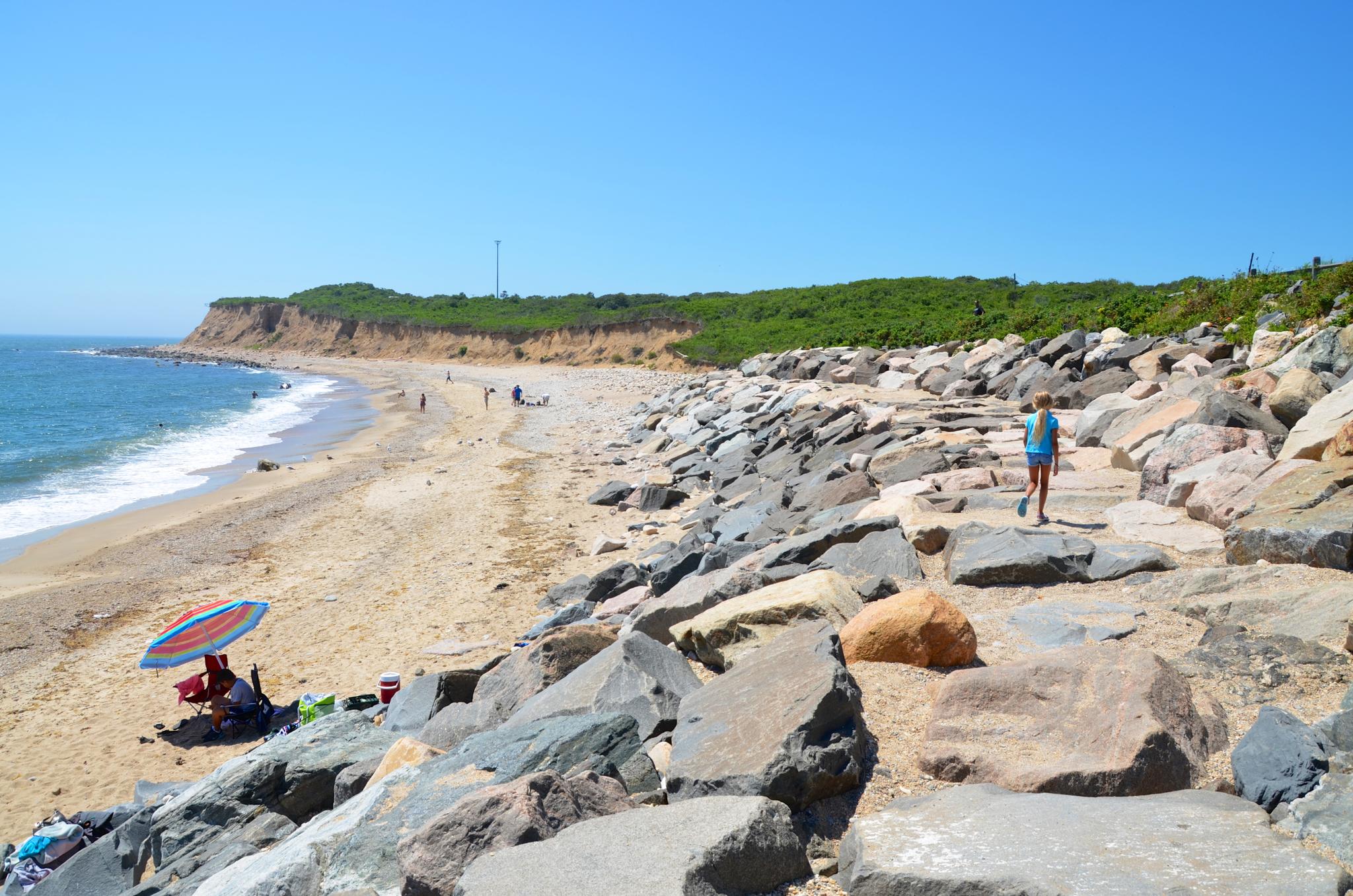 Sunny beach with a person walking on large rocks, others relaxing under an umbrella.