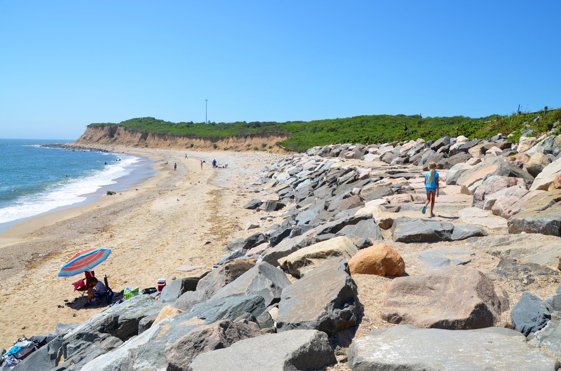 Sunny beach with a person walking on large rocks, others relaxing under an umbrella.