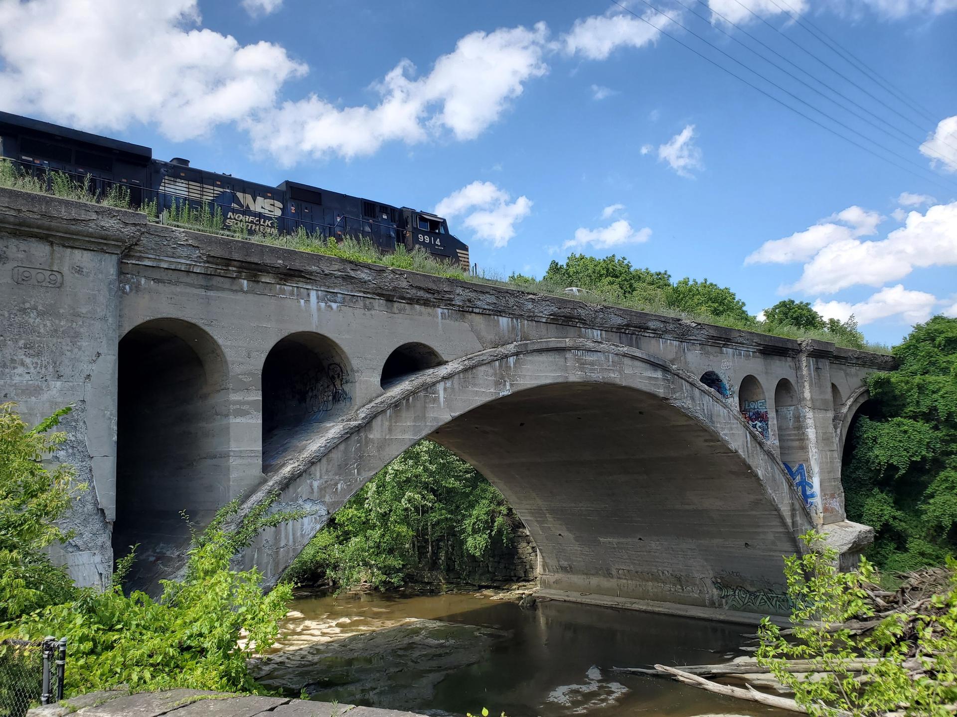Train crossing an arched concrete bridge over a river under a bright blue sky.