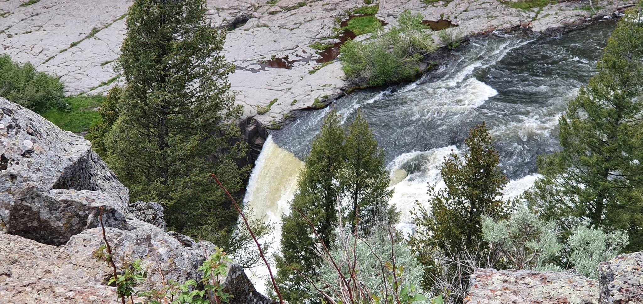 A river cascades over rocks, surrounded by trees and rocky banks.