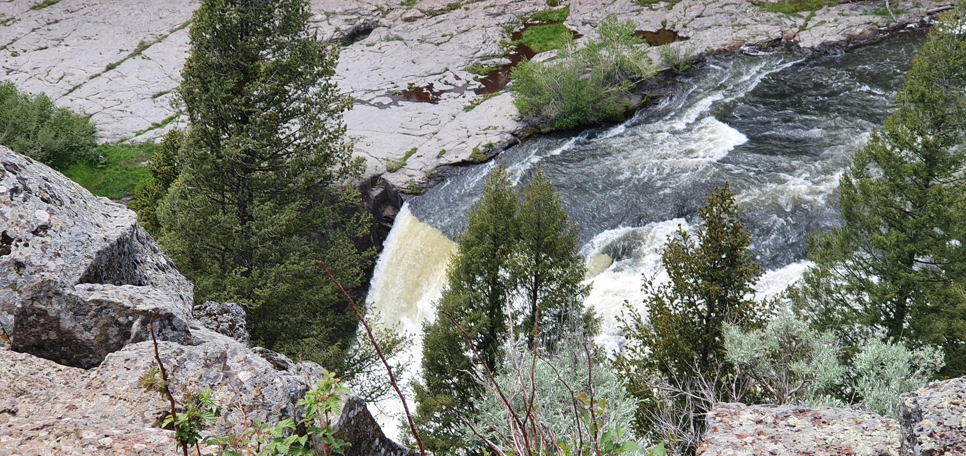 A river cascades over rocks, surrounded by trees and rocky banks.