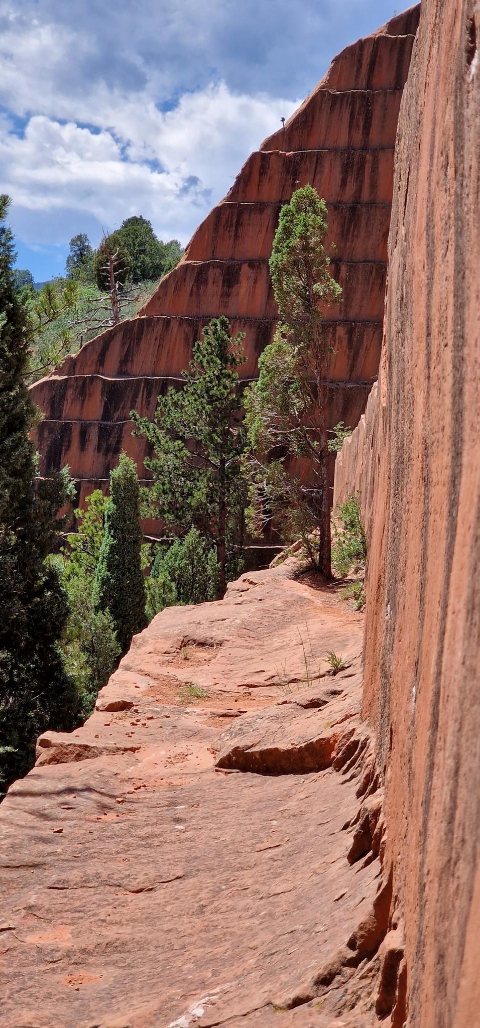 Narrow dirt path alongside a towering, layered red rock cliff with green trees.