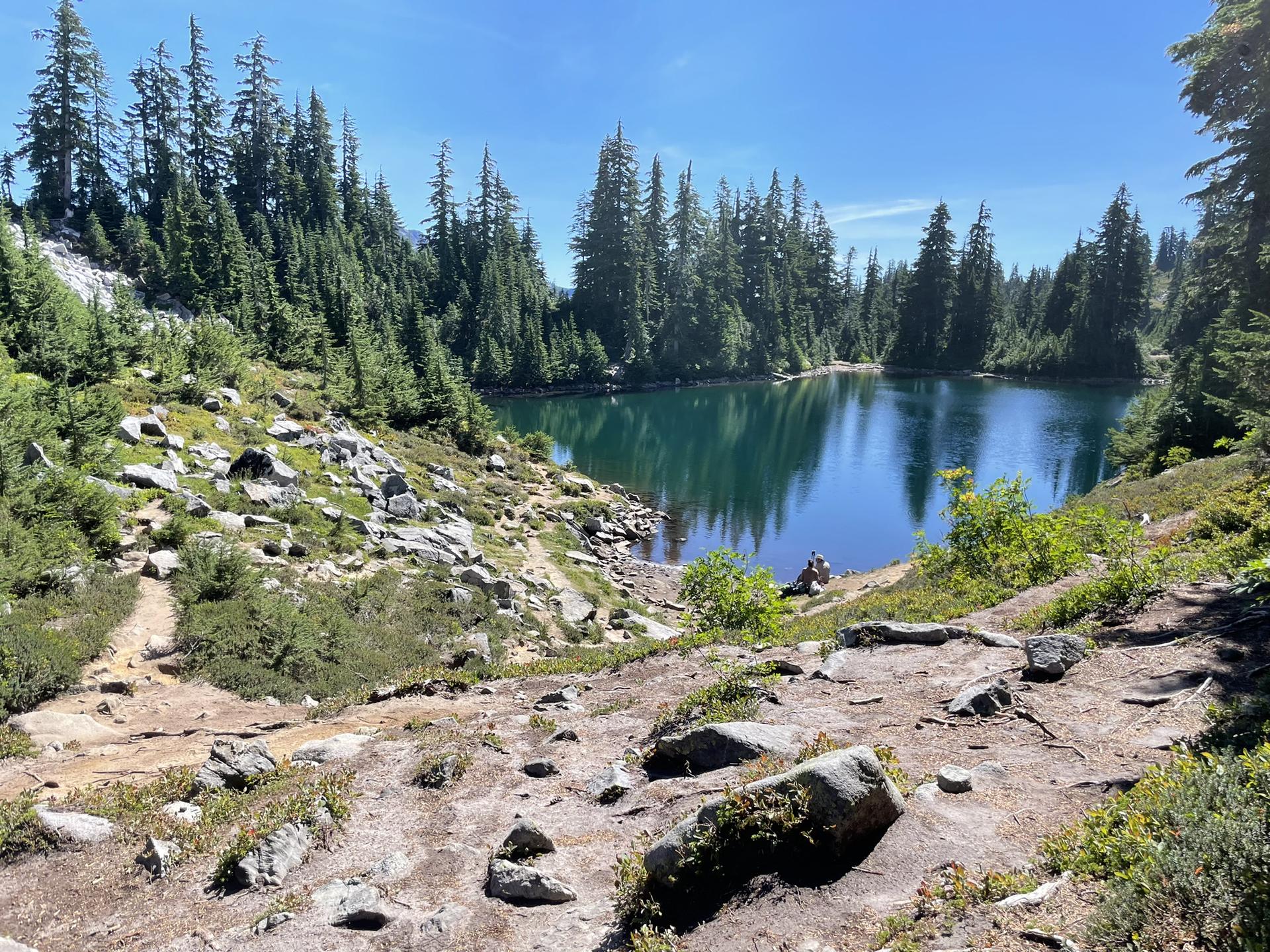 A serene blue lake nestled among evergreen trees and rocky terrain; a person sits.