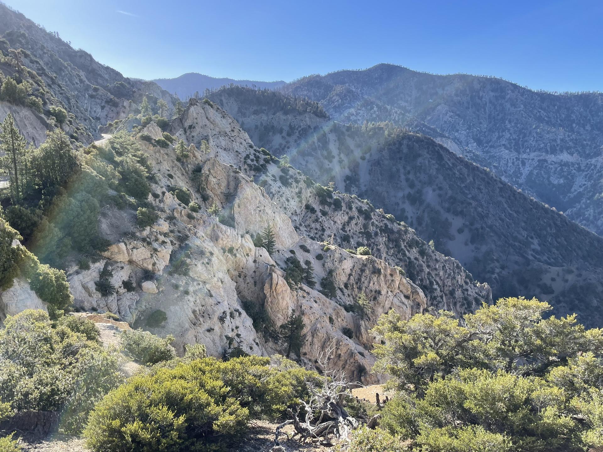 A vast mountain landscape with rocky cliffs, green trees, and bright sunlight.