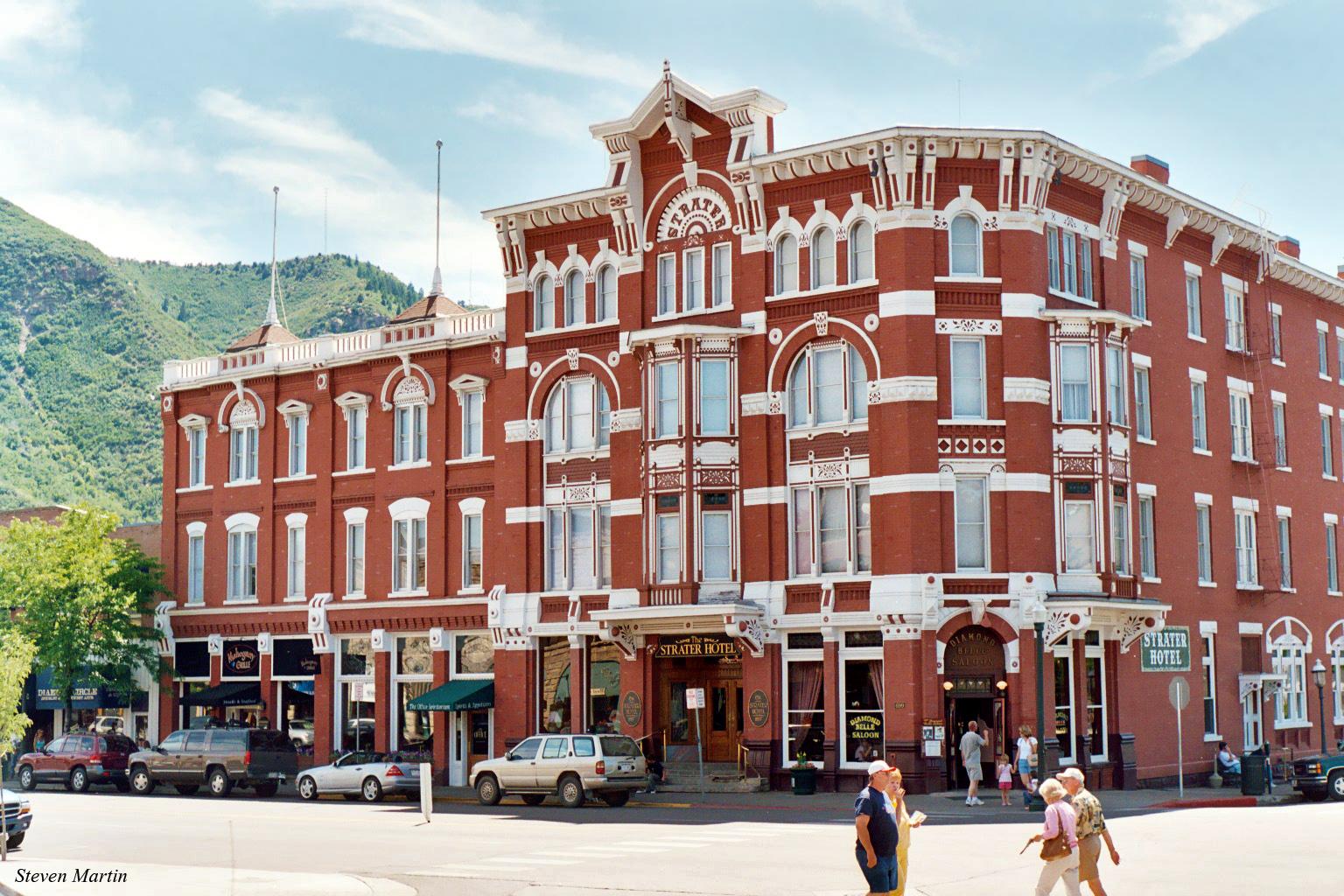 Ornate red brick building, people walking on street, green mountains, bright daylight.