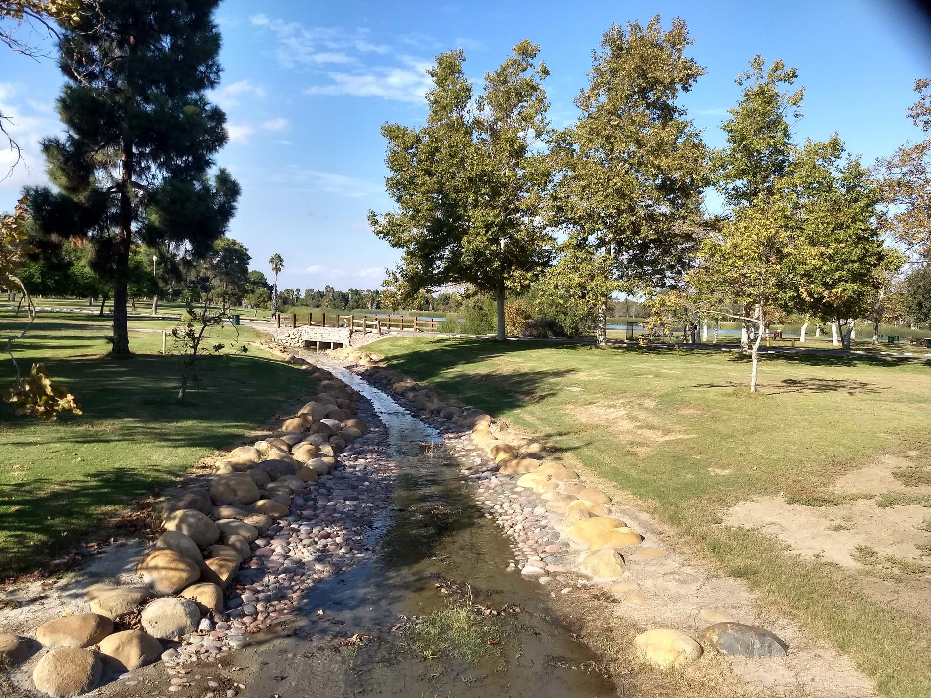 Small rocky stream flowing through a green park with trees and a distant bridge.