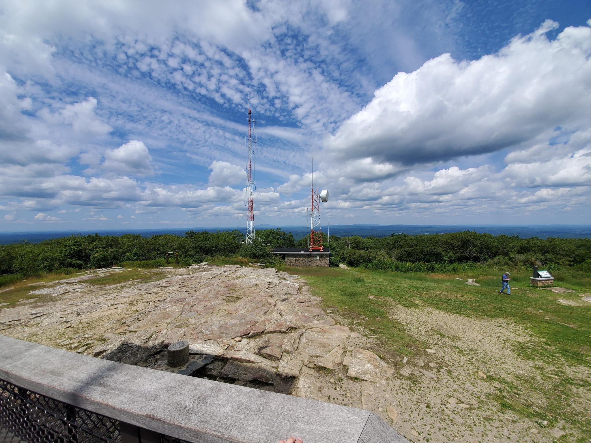 Panoramic view from a rocky summit with communication towers under a cloudy sky.