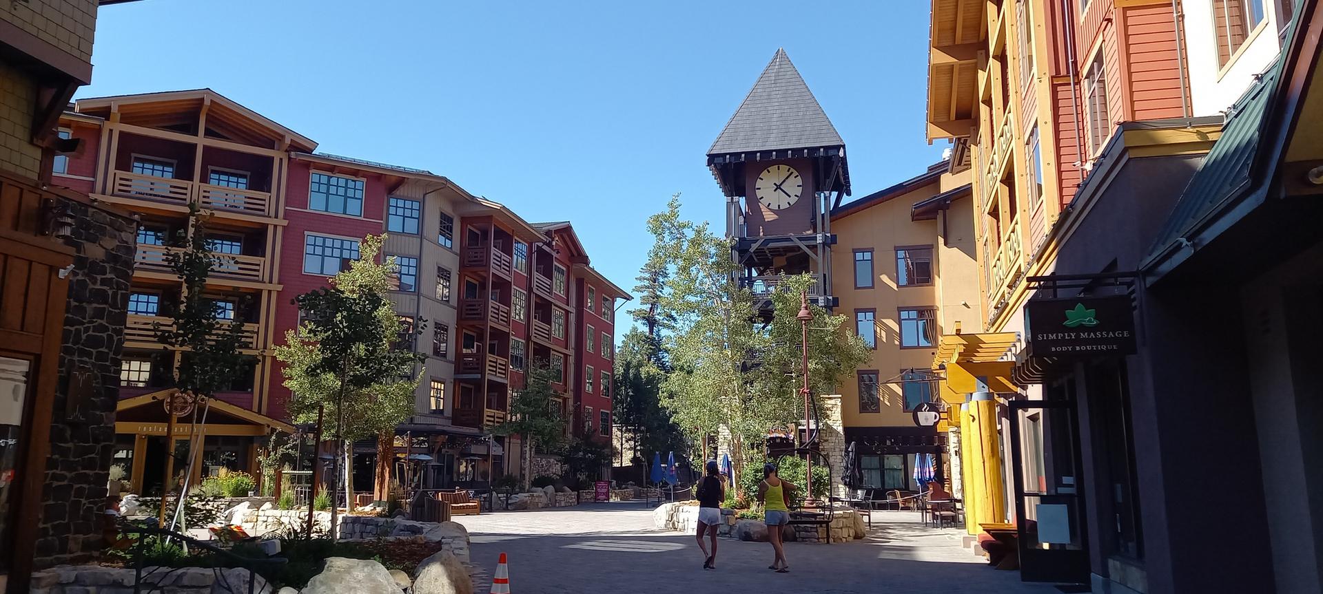 Sunny village square with multi-story buildings, a clock tower, and people walking.