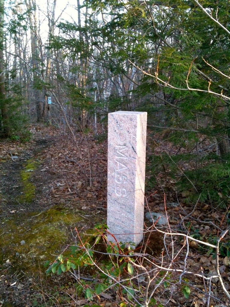 Tall stone marker with carved letters stands beside a dirt path in woods.