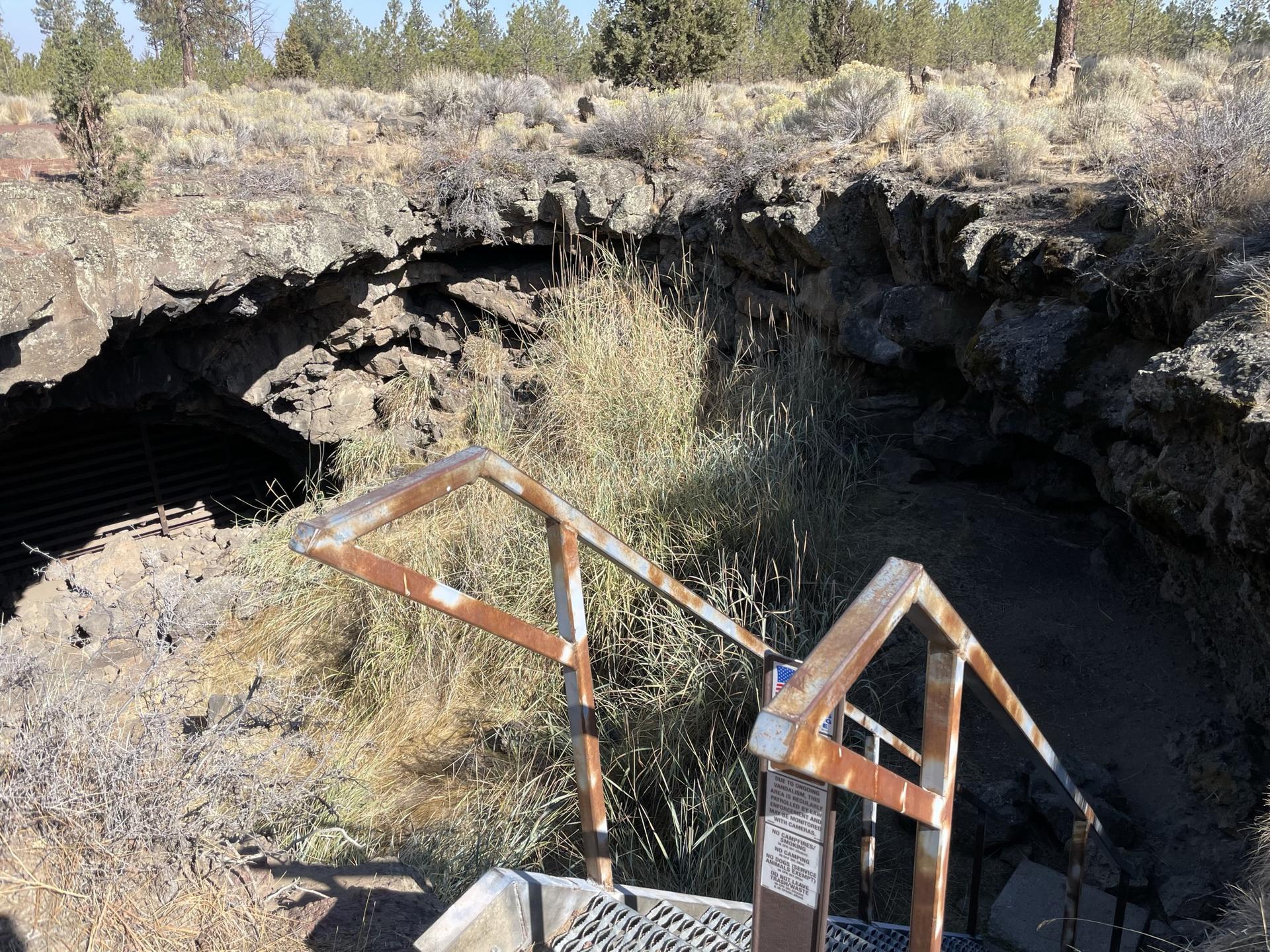 Rusty metal stairs leading into a dark cave entrance in rocky, dry terrain.