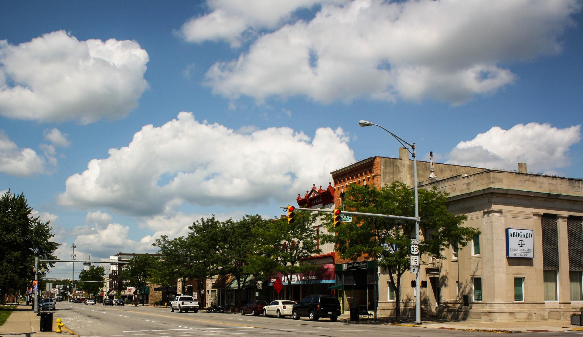 Sunny street scene with historic buildings, trees, and fluffy white clouds.