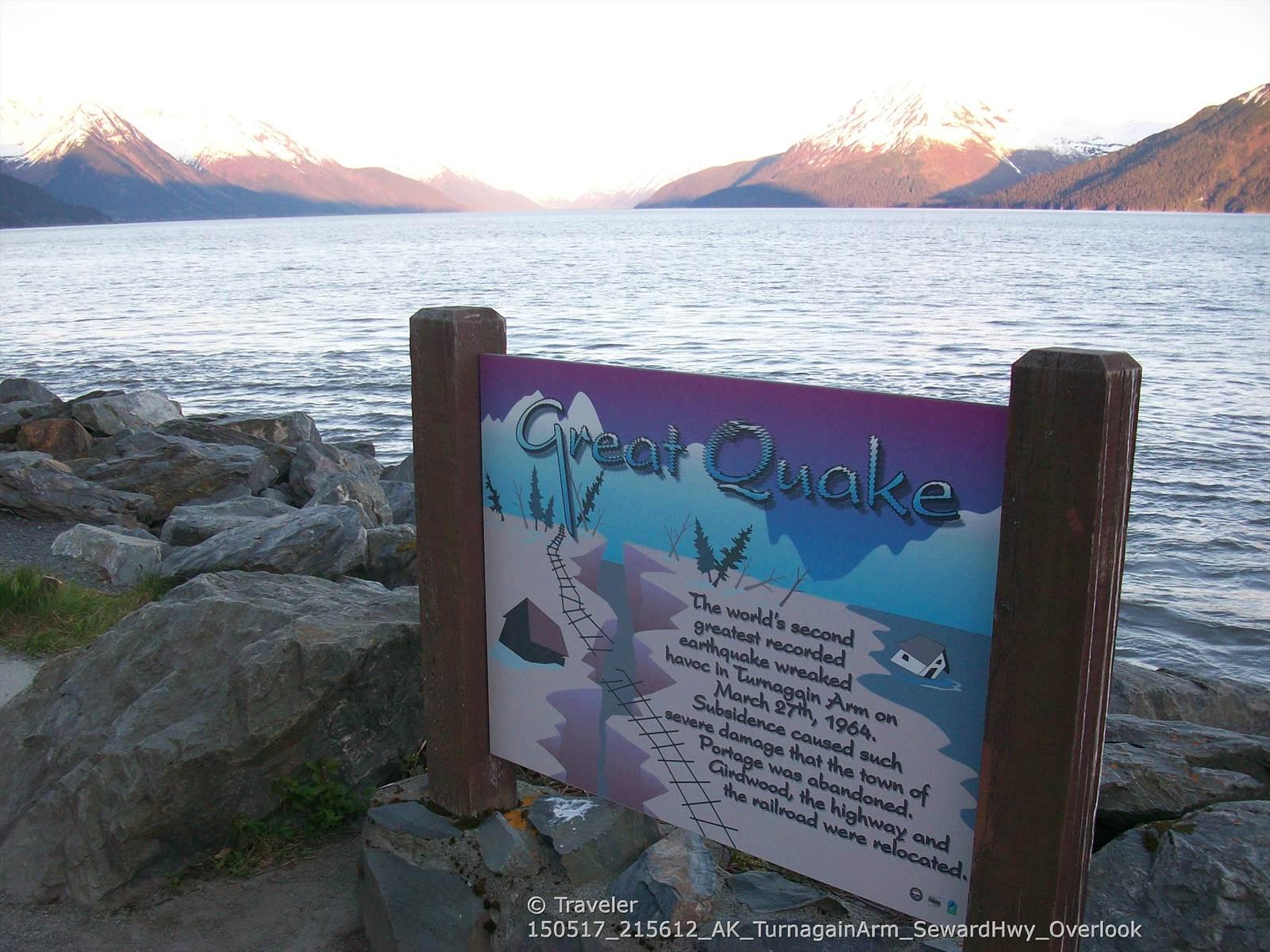Informational sign by a body of water with distant snow-capped mountains.