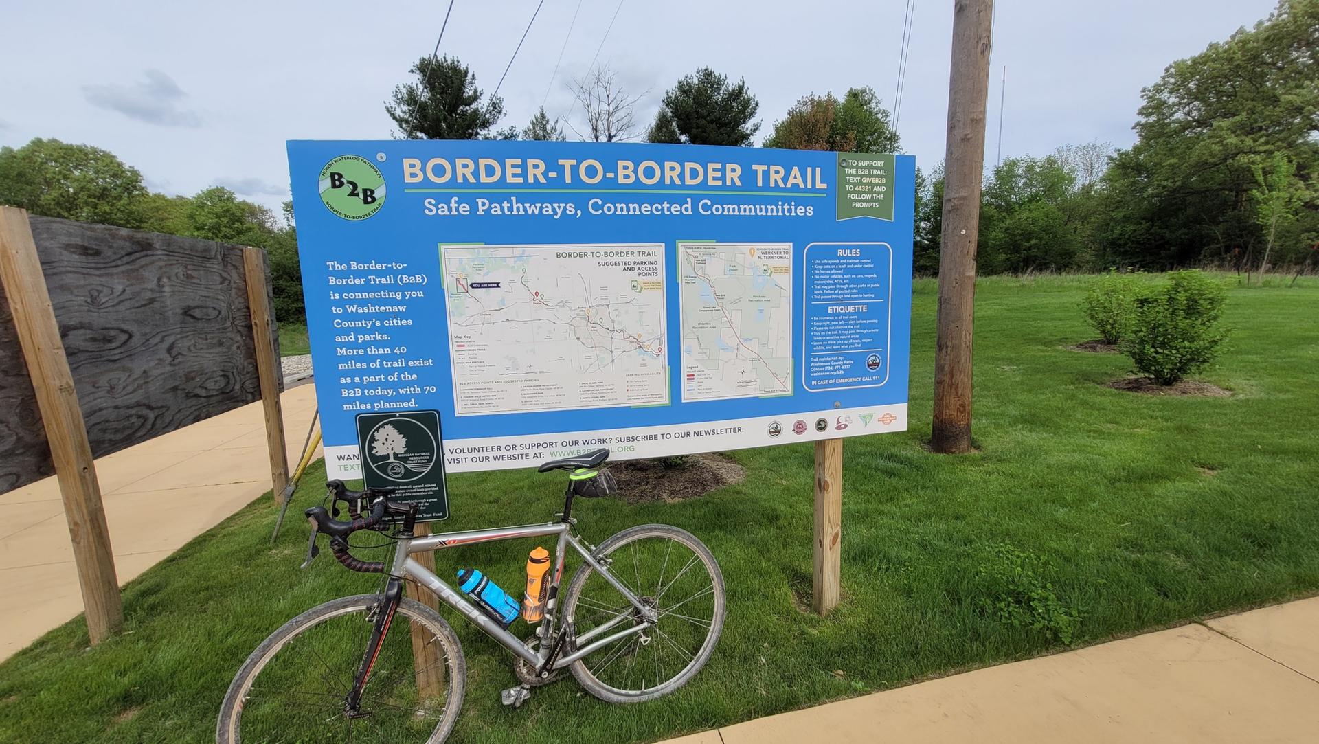 A bicycle rests beside a large blue trail information sign on a sunny day.