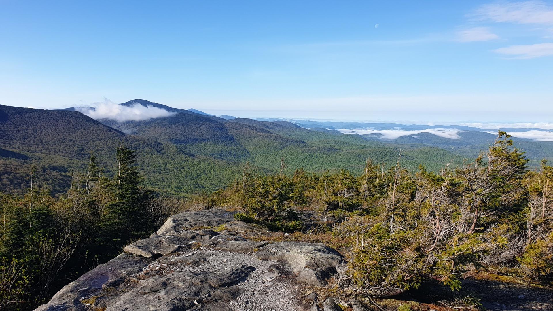 Rocky foreground overlooks vast green mountains under a clear blue sky.