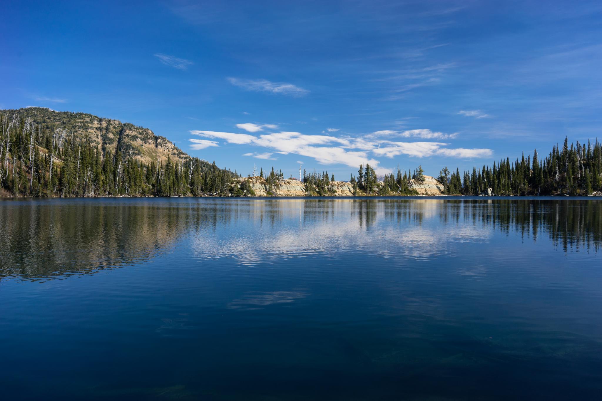 Calm lake reflecting forested hills under a bright blue sky with clouds.