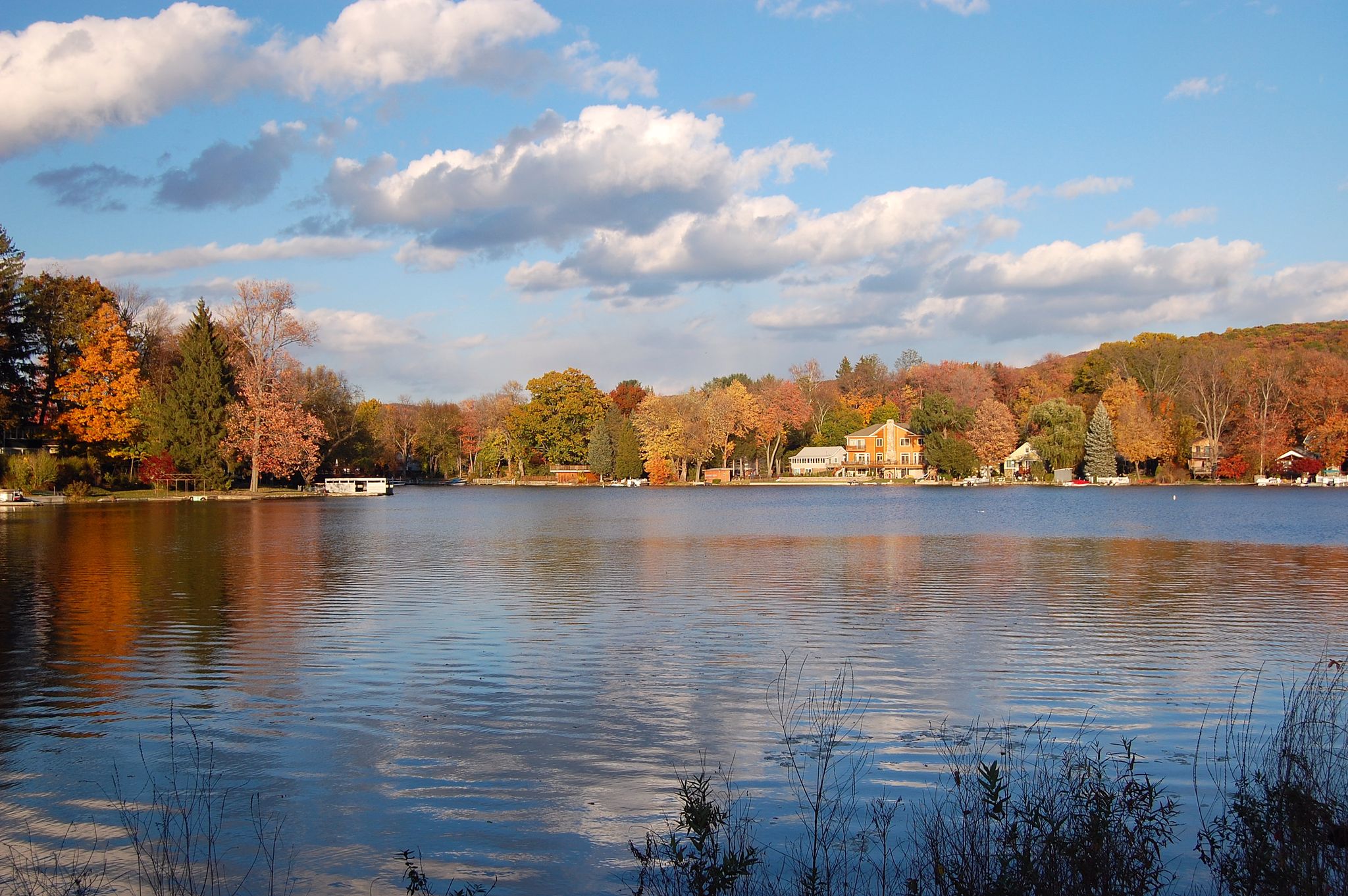 A wide lake reflects colorful autumn trees and houses under a bright sky. - Monksville Reservoir Loop