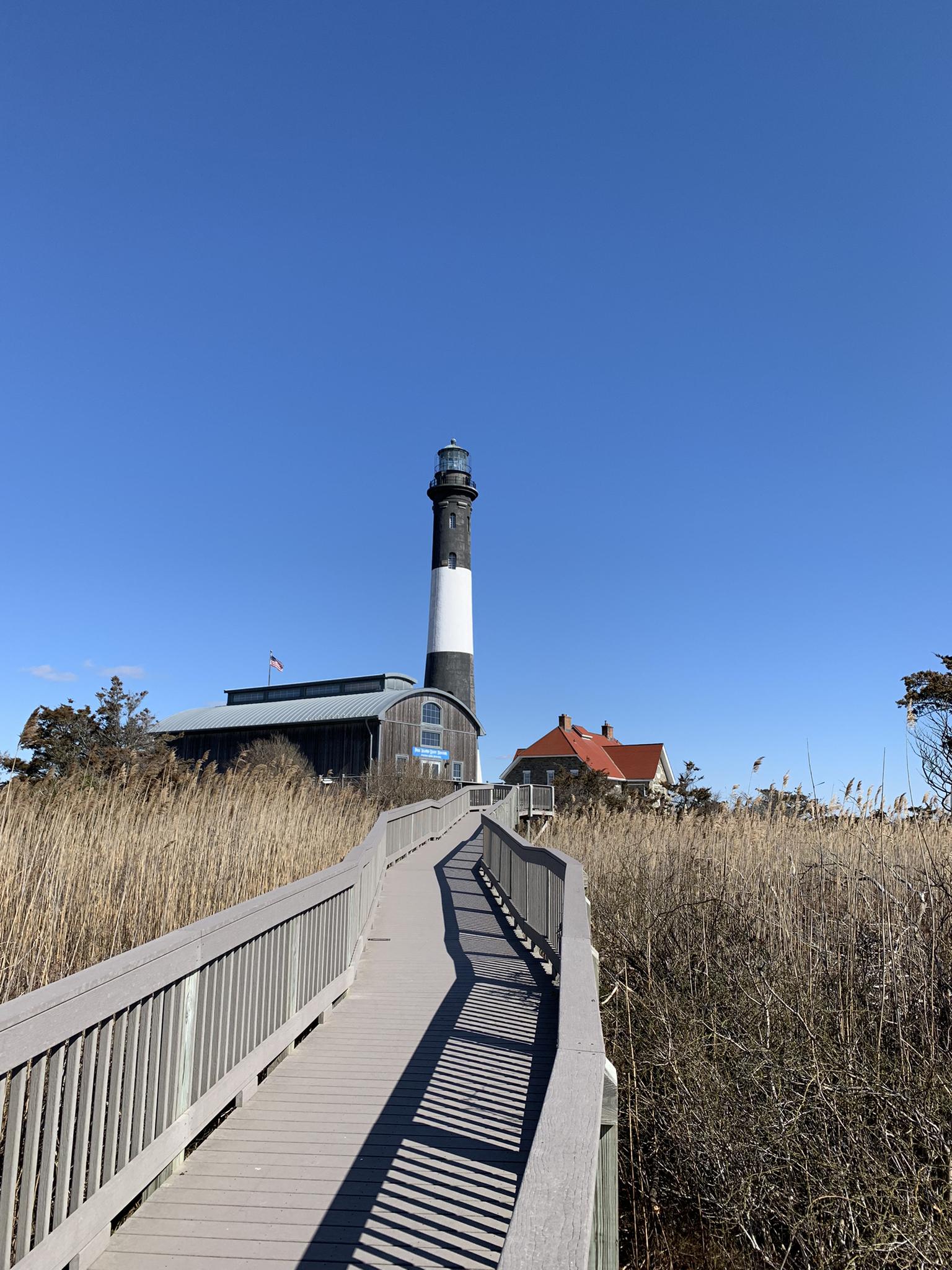 Wooden boardwalk leads to a tall black and white lighthouse under a clear sky.