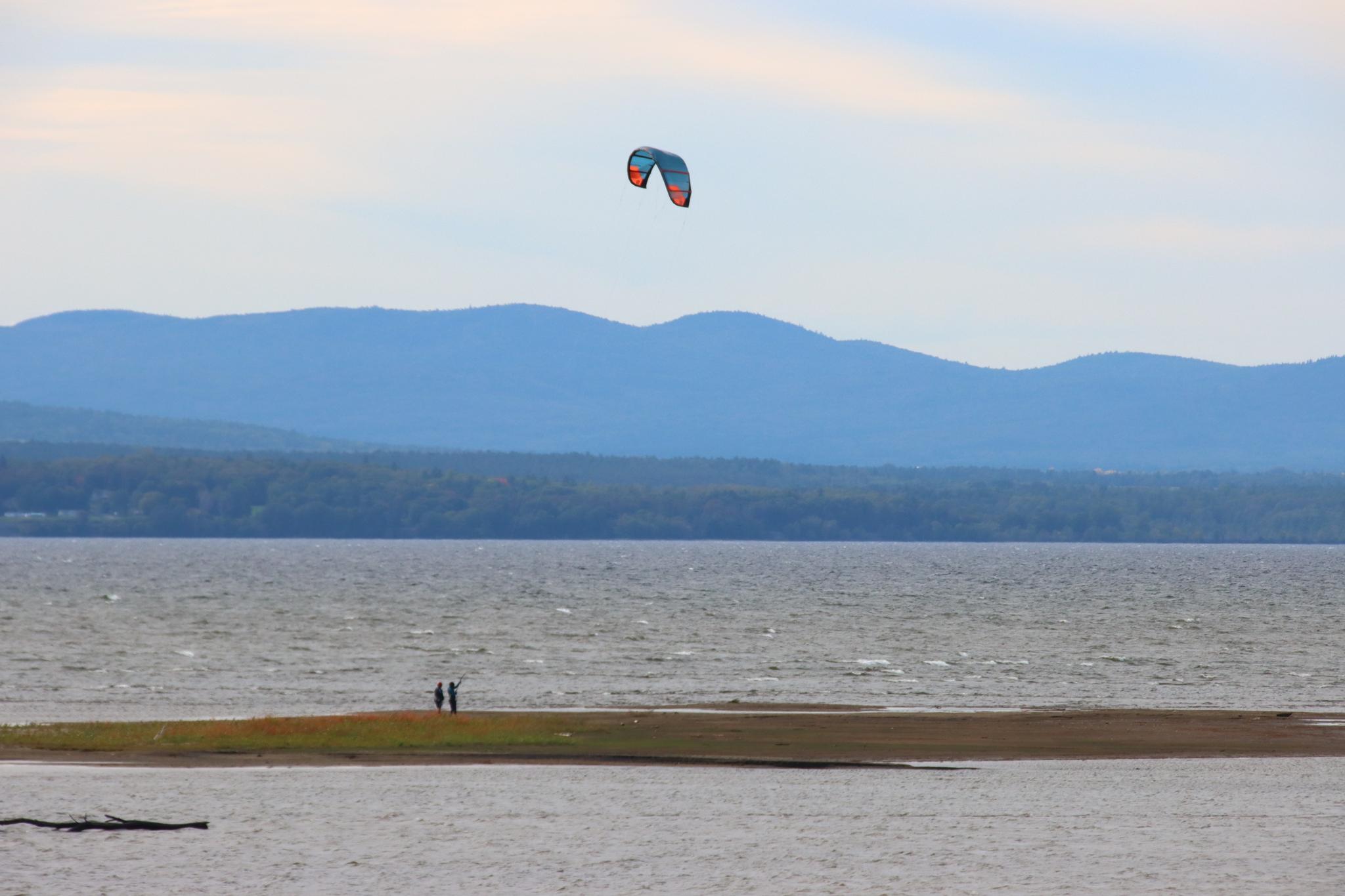A kitesurfer glides over sparkling water with distant mountains under a cloudy sky.