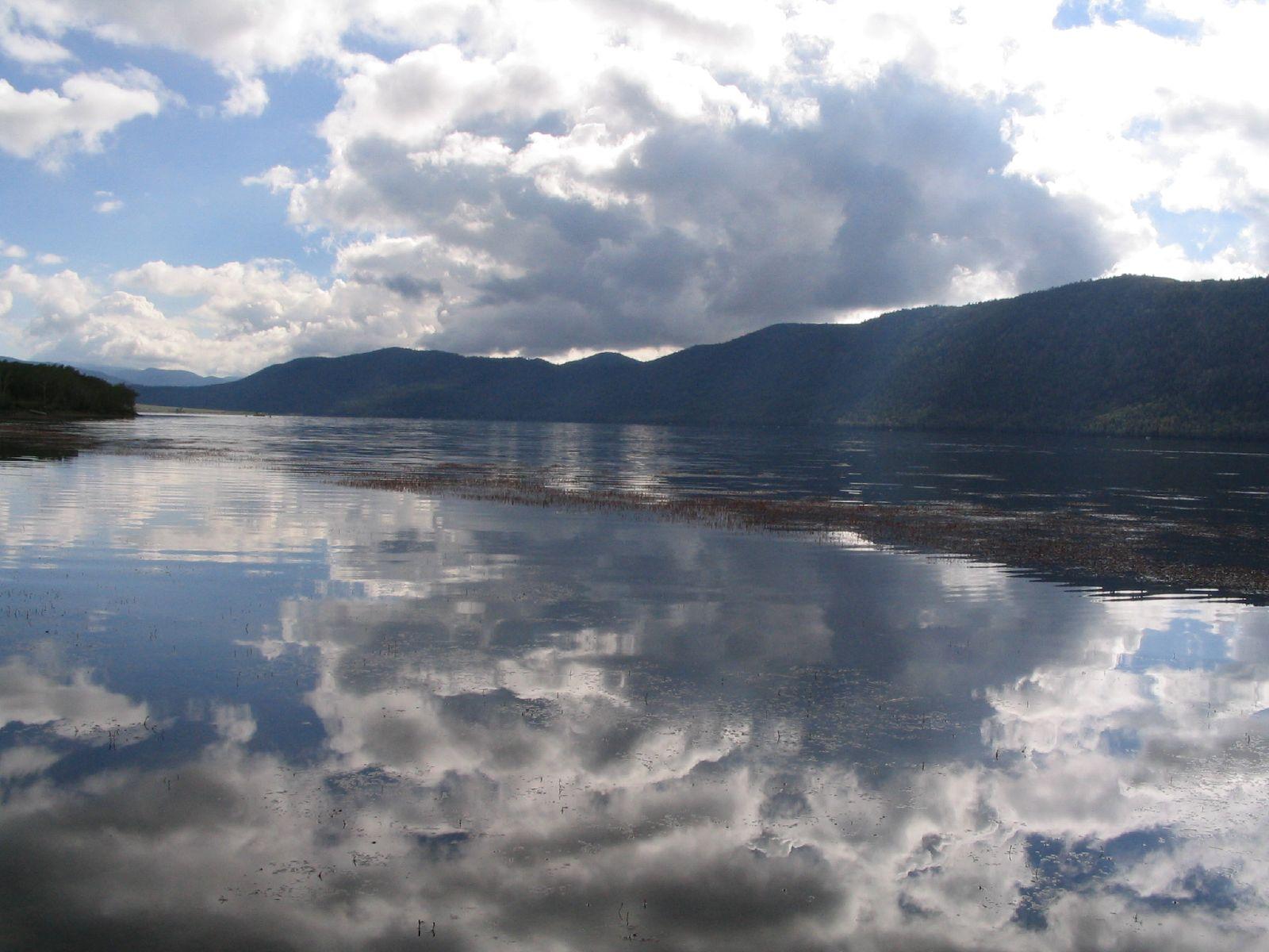 A wide lake reflects a cloudy sky, with dark mountains in the distance.
